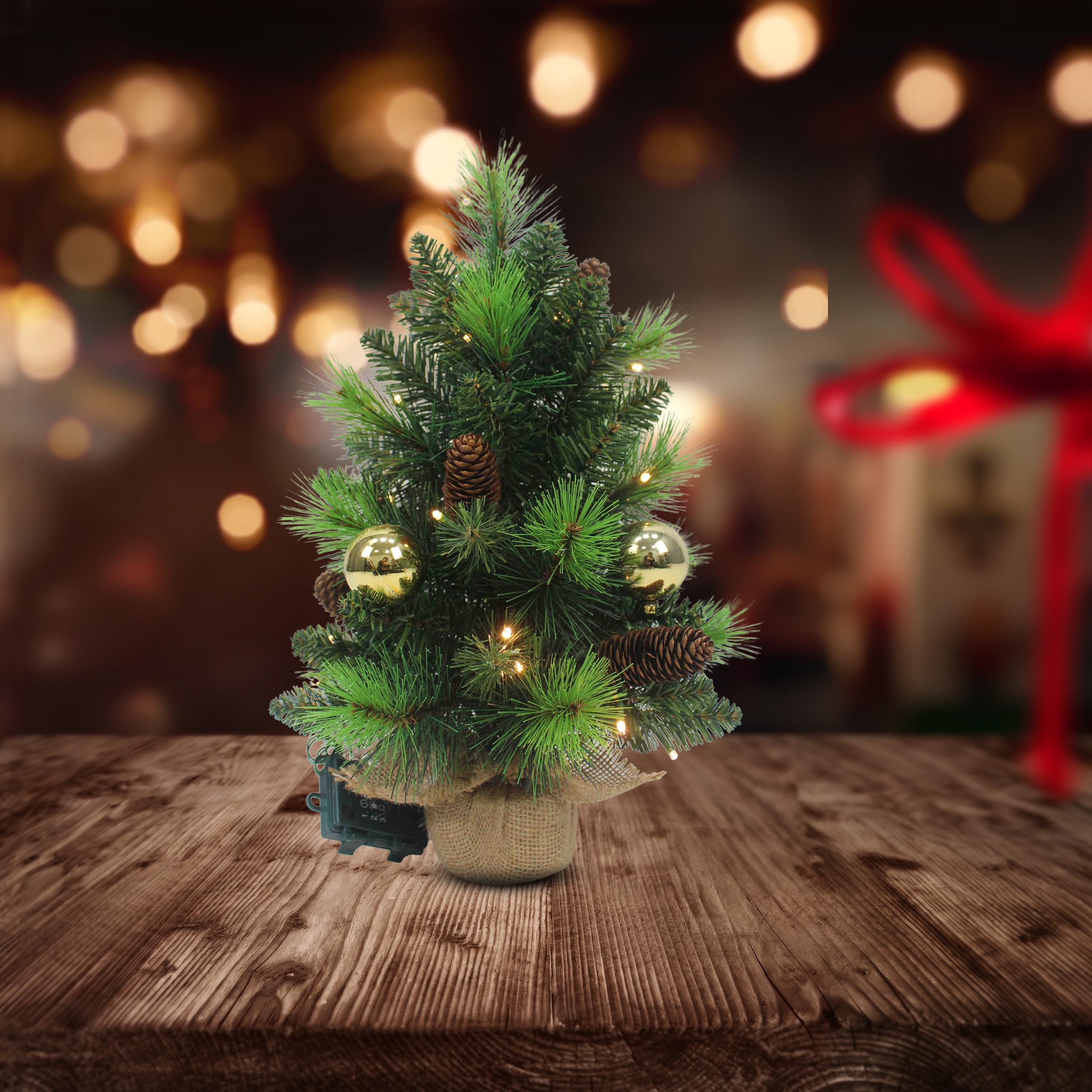 Kleiner künstlicher Weihnachtsbaum auf einem Holztisch. Mit Lichtern und Ornamenten geschmückt.