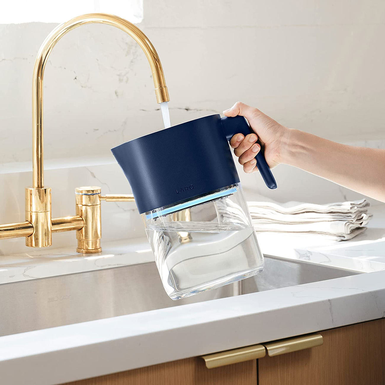 Persona che versa acqua in una brocca filtrante blu in un lavandino con un rubinetto dorato.