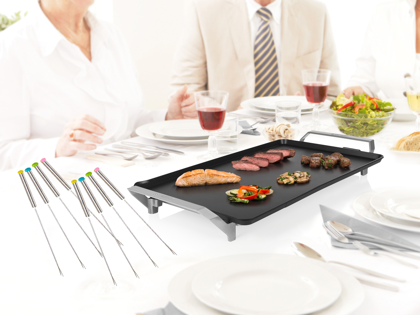 Ein Grill mit gekochtem Essen und Personen im Hintergrund. Fonduegabeln sind vorne.