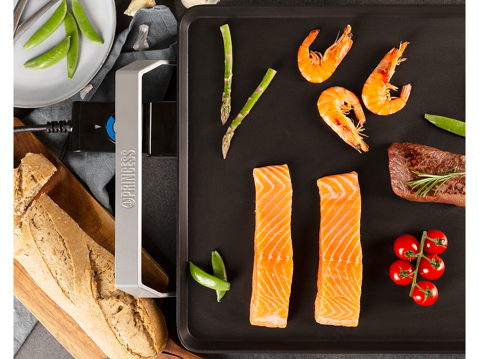 Draufsicht auf eine Grillplatte mit Essen: Garnelen, Lachs, Spargel, Erbsen und Tomaten.