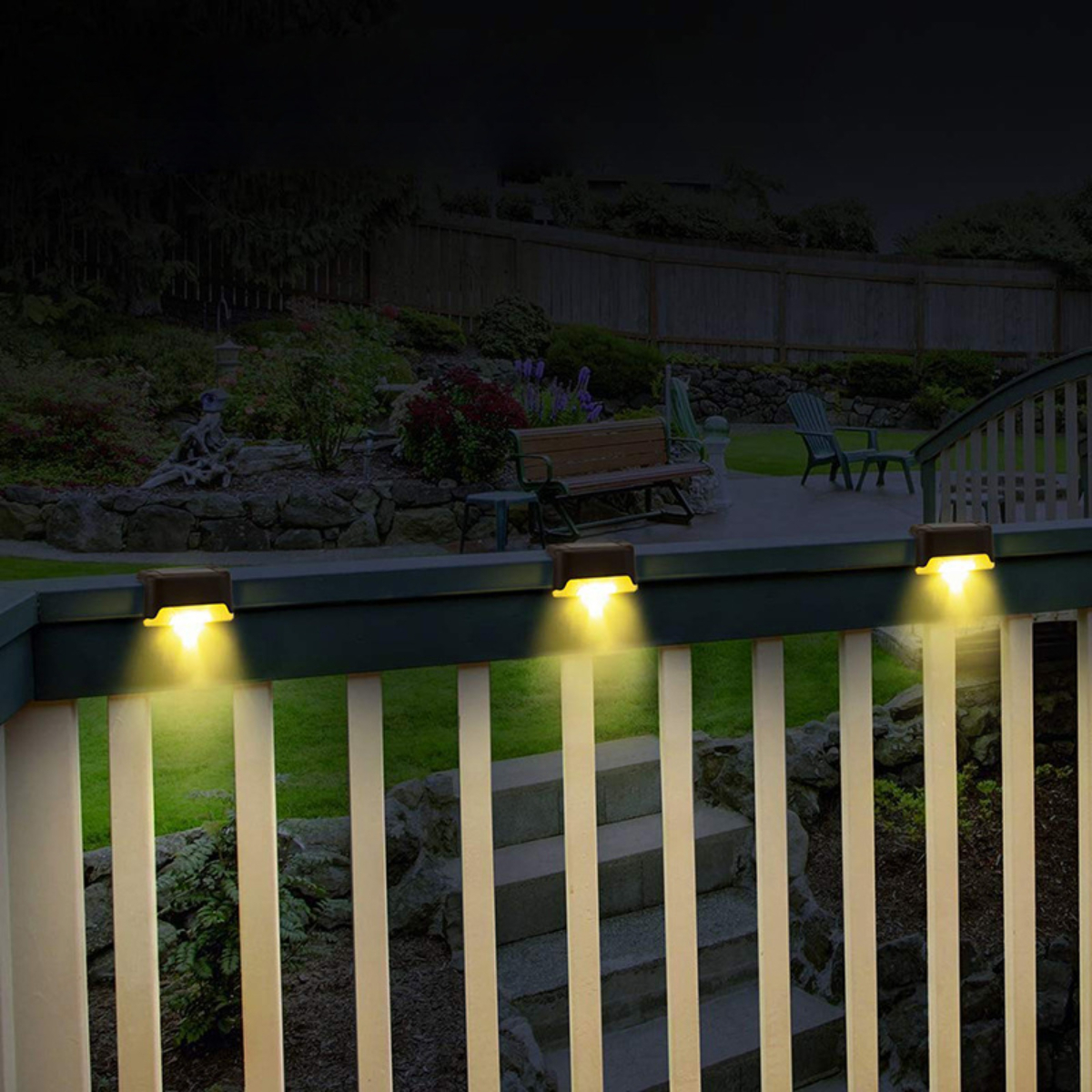 Drei Außenleuchten an einem Geländer, die nachts einen Garten beleuchten. Die Lichter sind gelb.