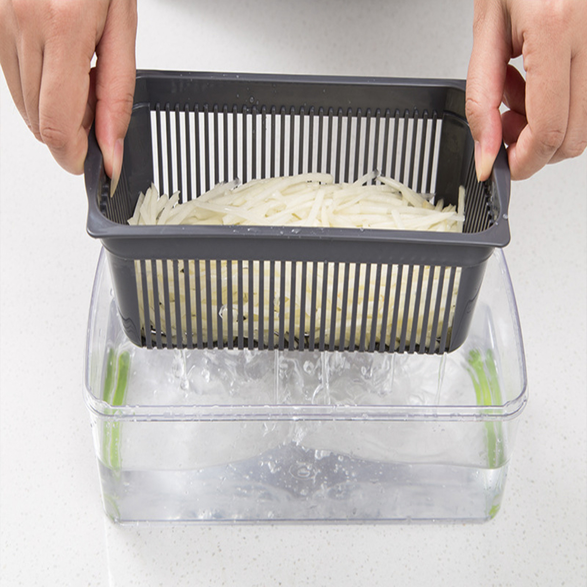 Manos sosteniendo un colador gris con papas en rodajas sobre un recipiente con agua.