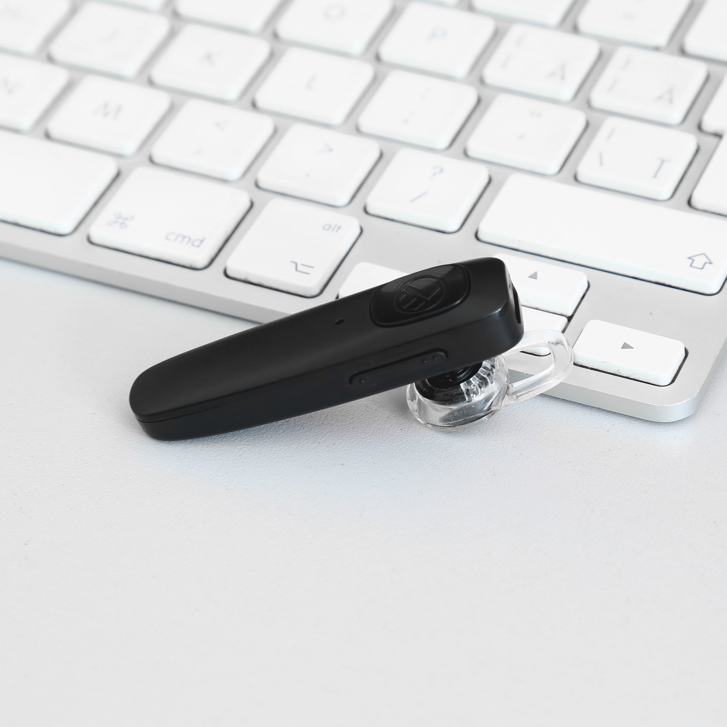 Schwarzes Bluetooth-Headset auf einer weißen Tastatur. Durchsichtiges Ohrstück befestigt.