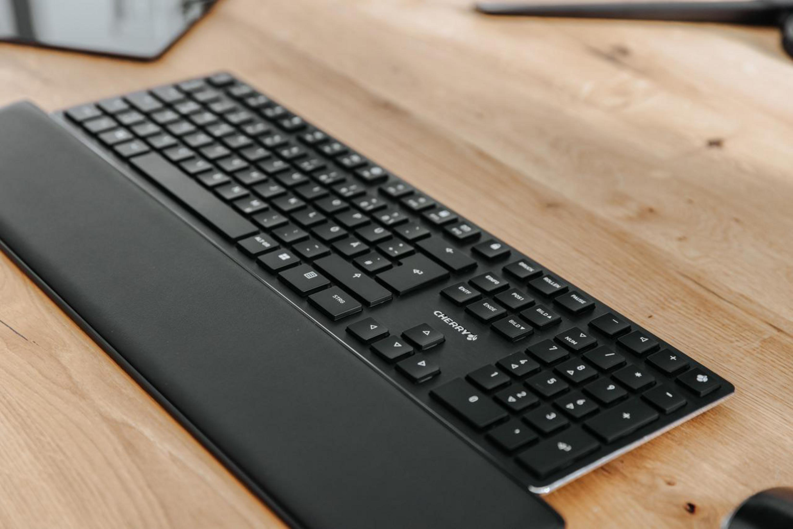 Czarna klawiatura z podpórką pod nadgarstki na drewnianym stole. Widoczna jest marka 'Cherry'.