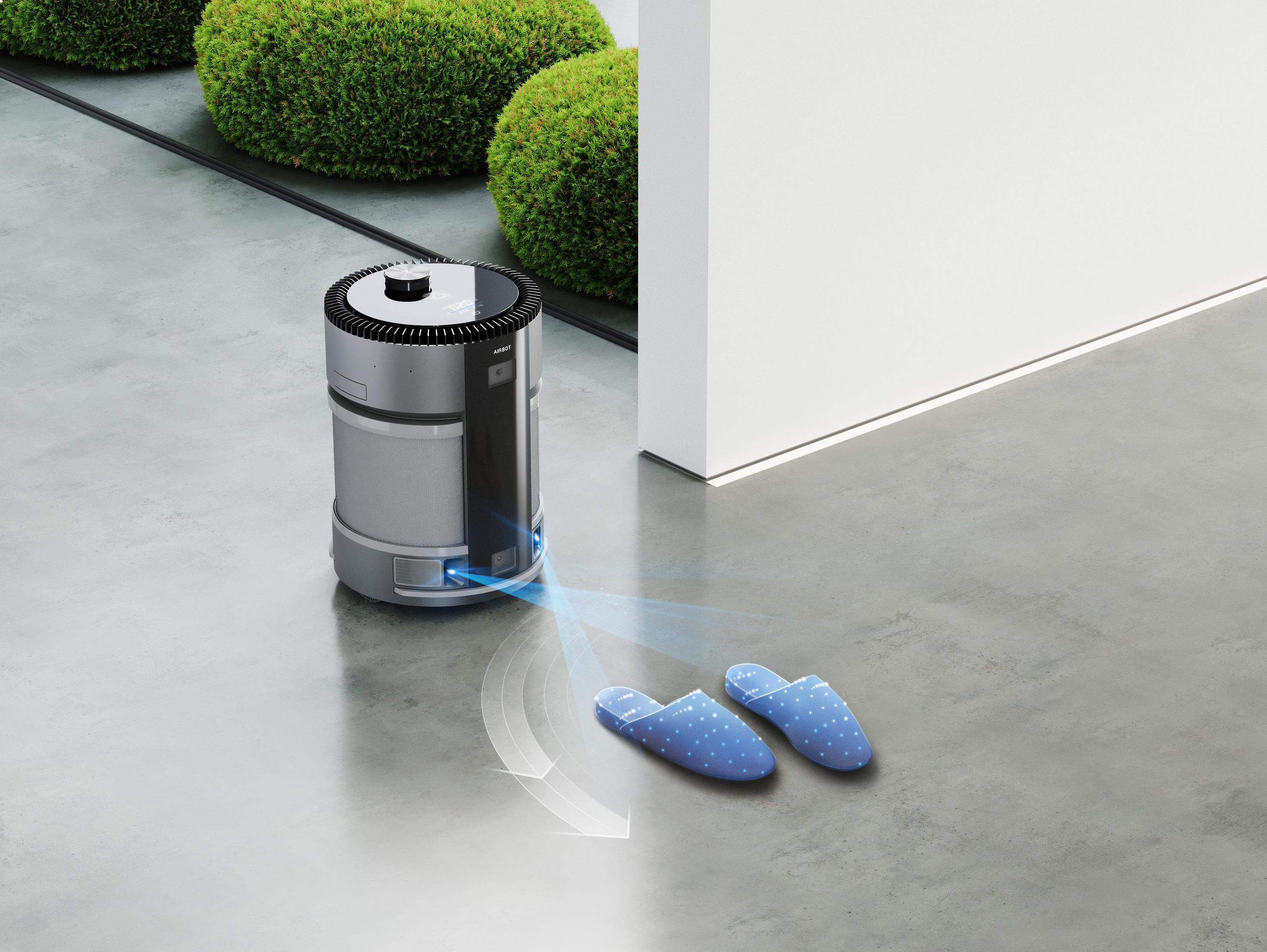 Ein silberner und schwarzer Roboter-Staubsauger in der Nähe blauer Hausschuhe auf grauem Boden.