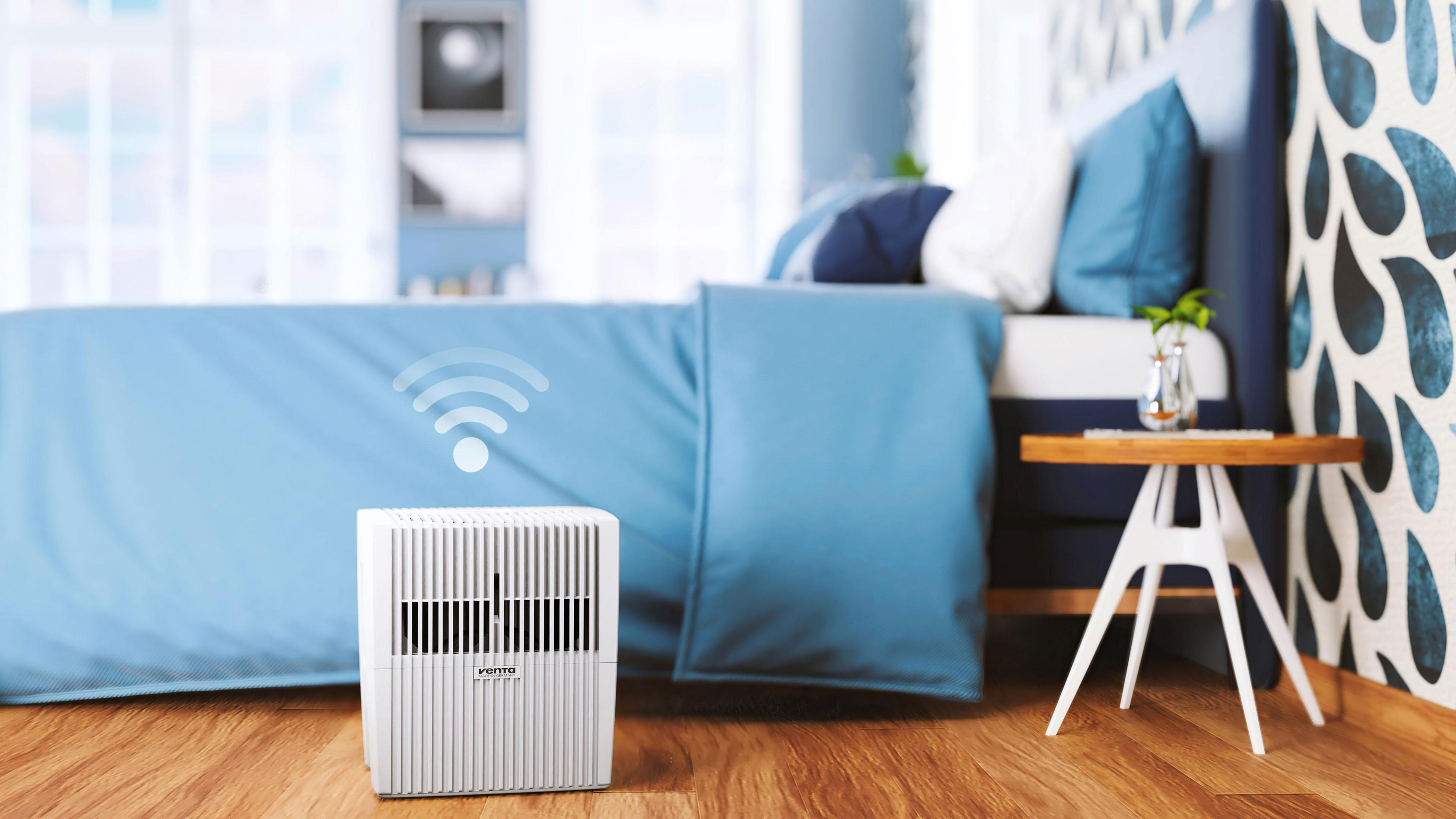 Biały nawilżacz powietrza na drewnianej podłodze, obok łóżka z niebieską pościelą.