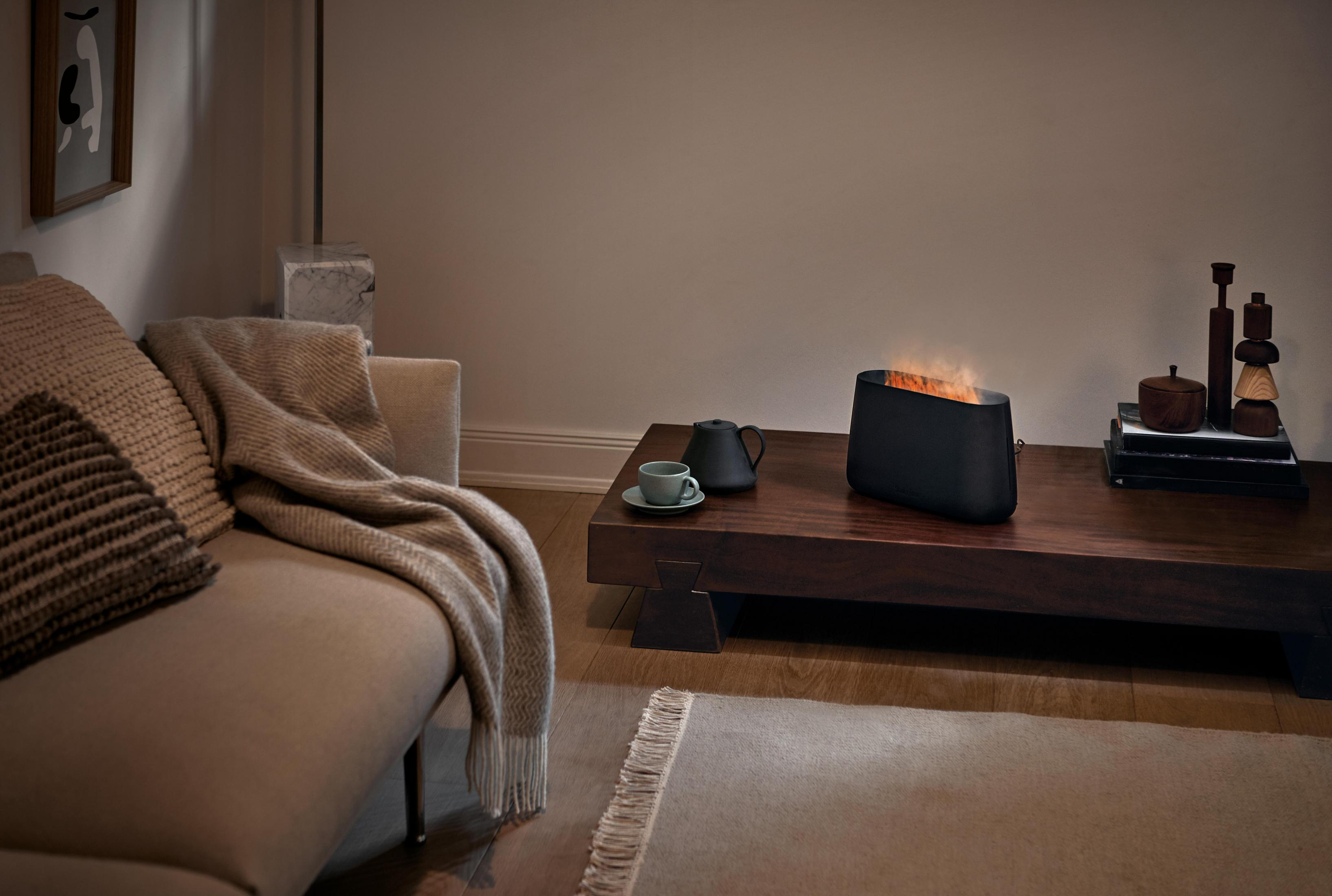 Czarny kominek dekoracyjny na stole. Beżowa sofa, dywan i stolik kawowy obecne.