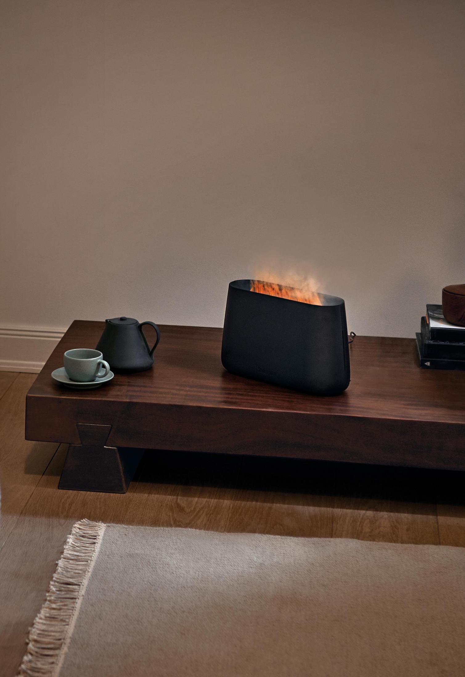 Czarny prostokątny kominek na drewnianym stole. Z boku filiżanka i czajnik.