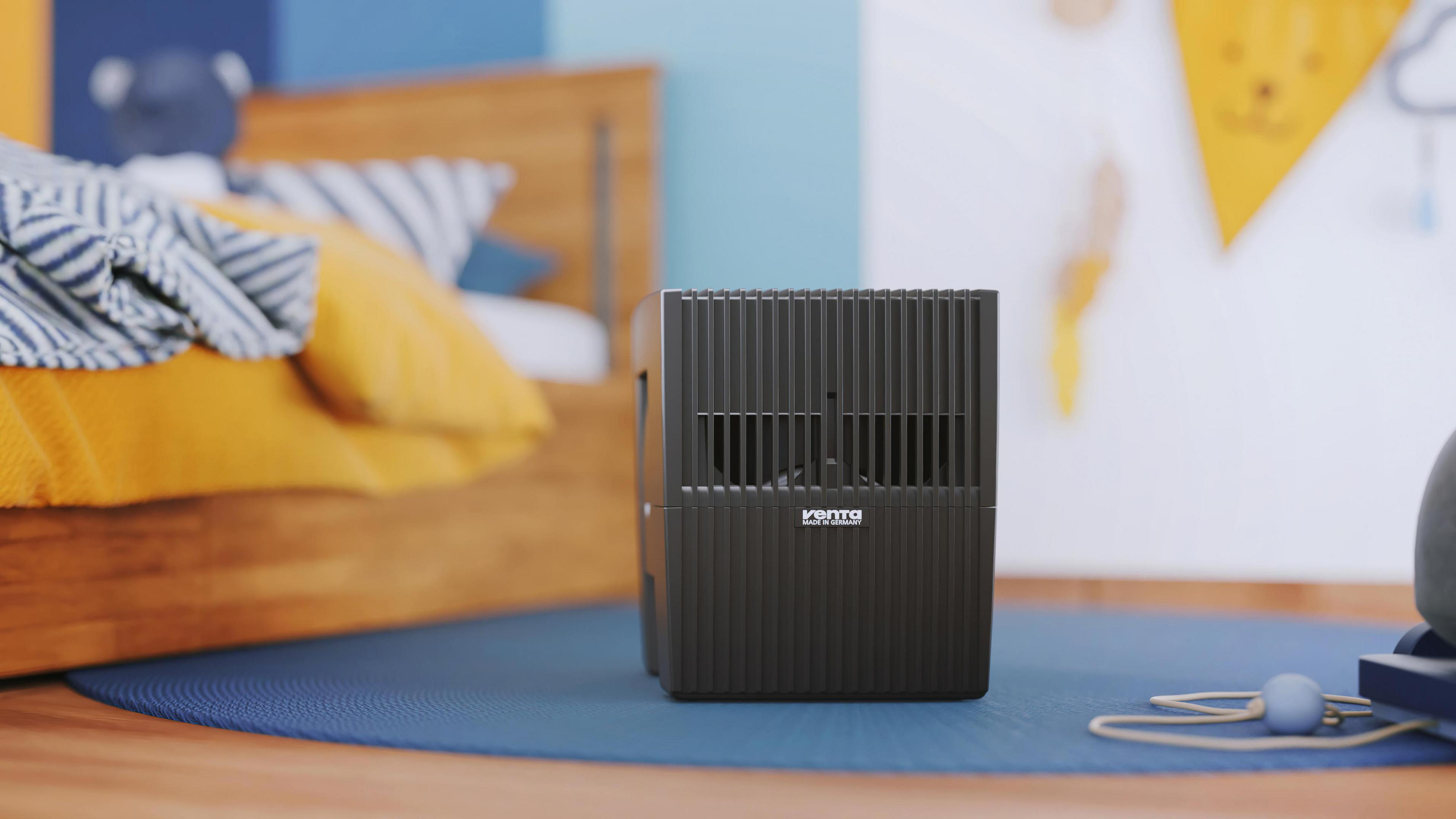 A black air purifier in a child's bedroom. It sits on a blue rug near a bed and a wall decoration.