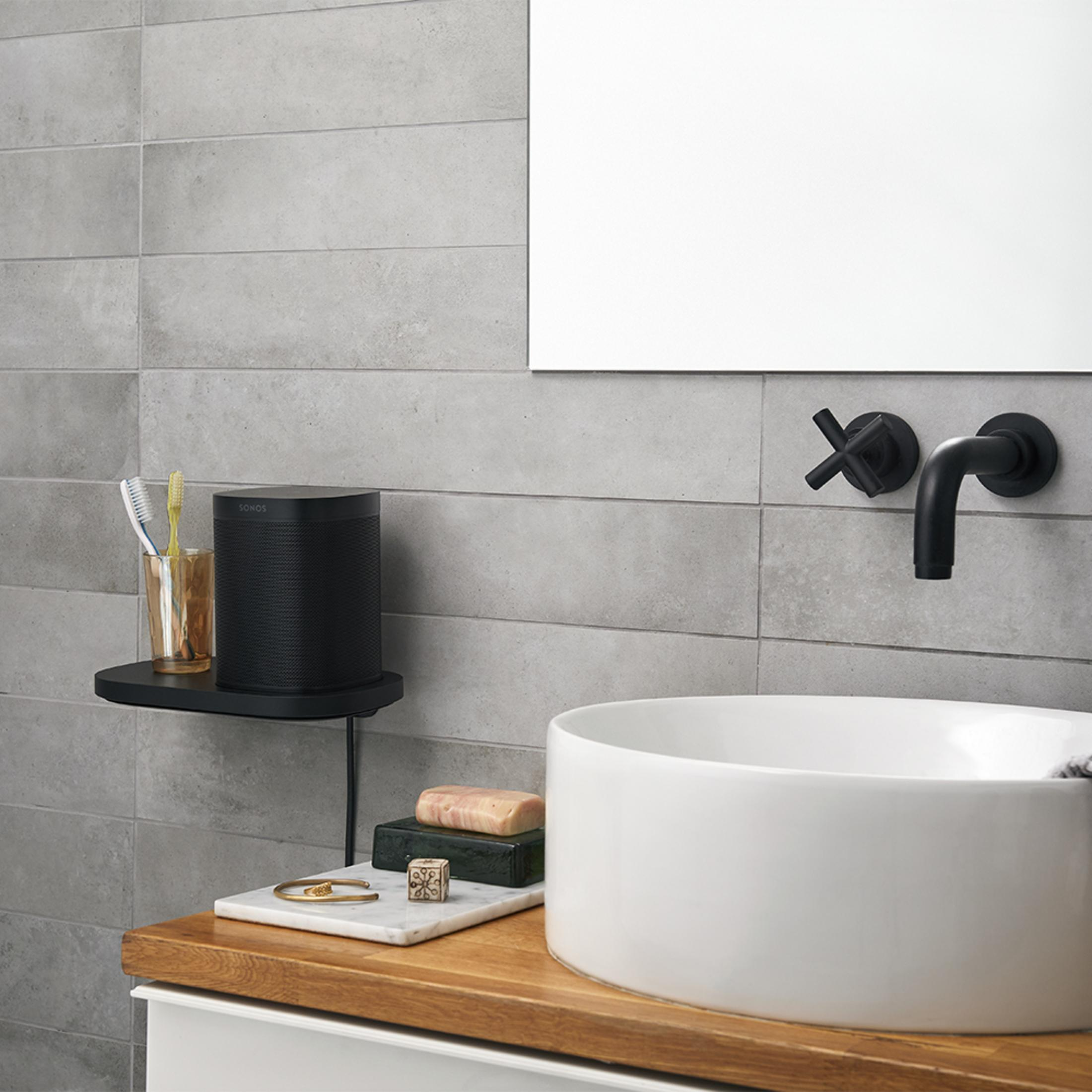 Escena de baño con un altavoz negro en un estante, lavabo y espejo. Superficie de madera, baldosas grises.