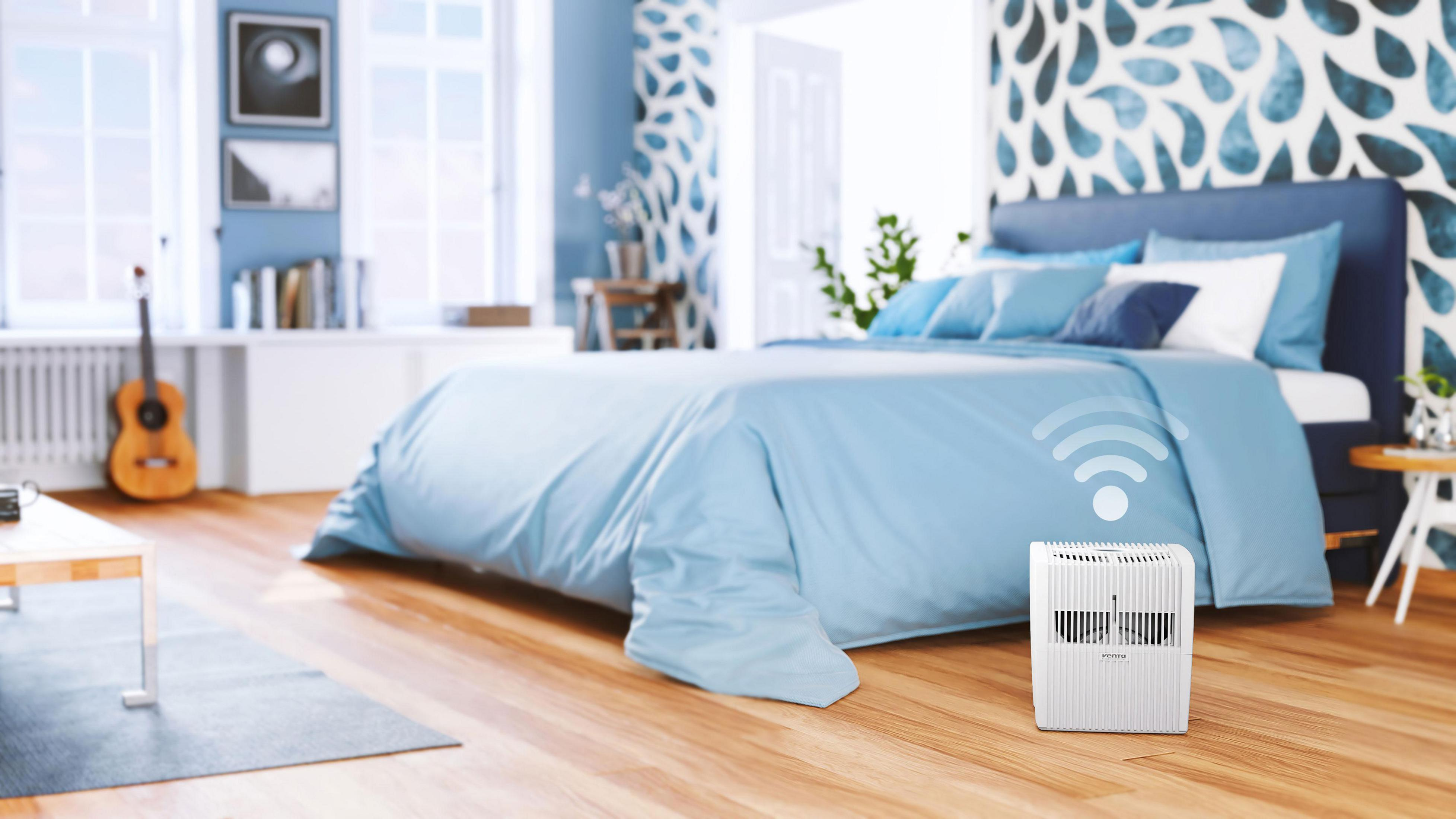 Biały oczyszczacz powietrza obok łóżka w niebieskiej sypialni, z wyświetlanym symbolem Wi-Fi.