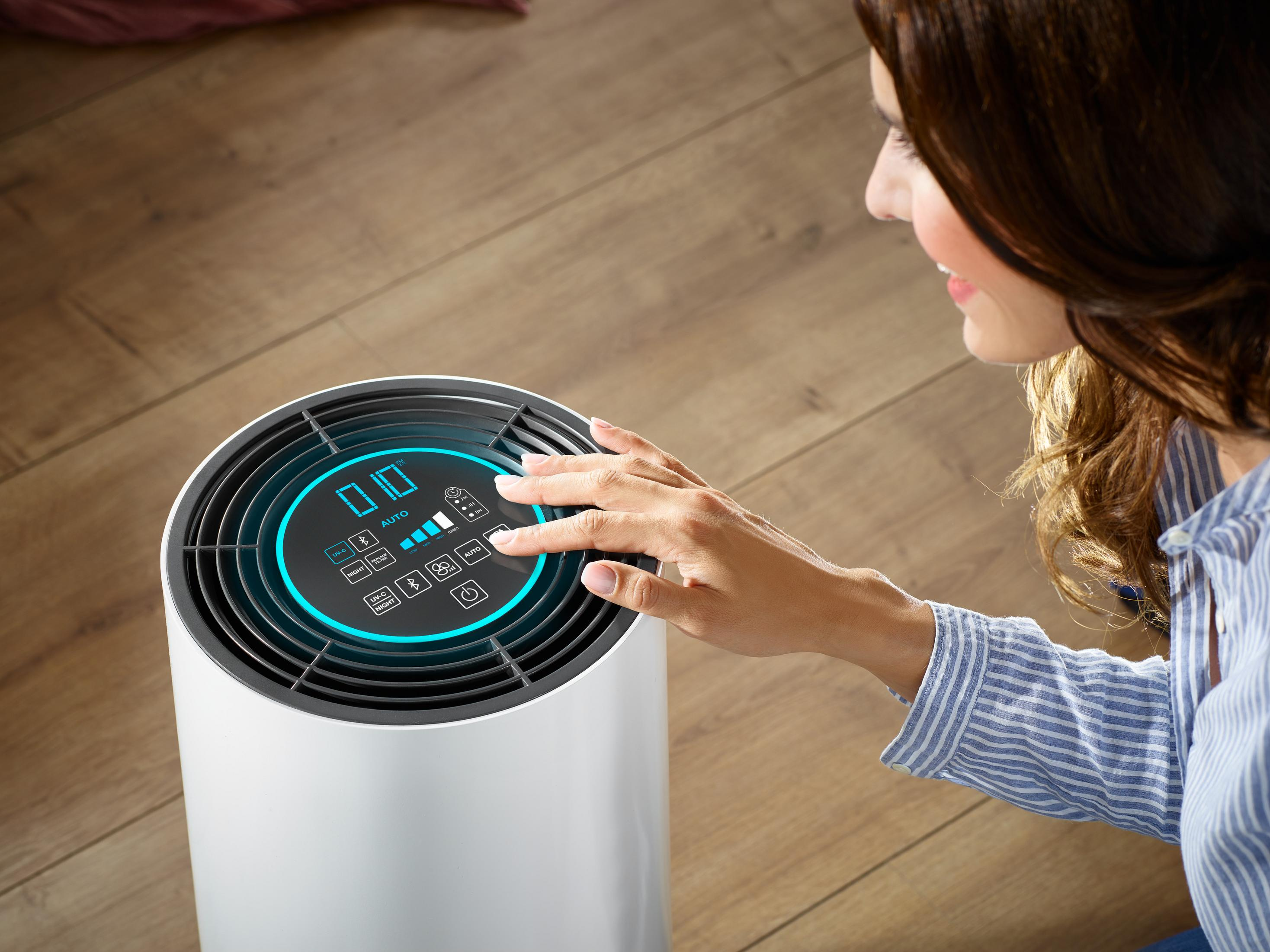 Die Hand einer Frau berührt das Bedienfeld eines Luftreinigers. Der Reiniger ist weiß und zylindrisch.