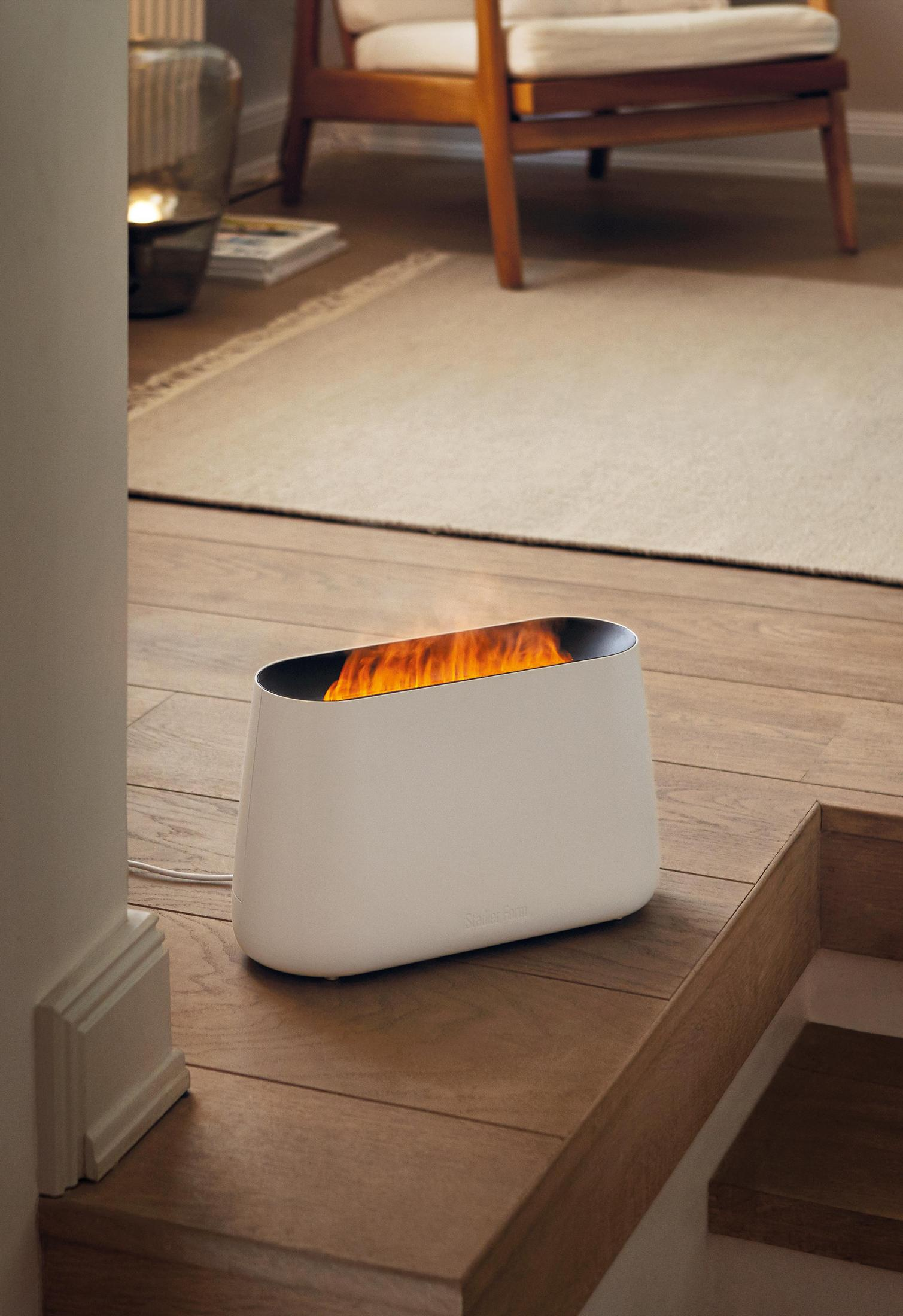 Biały grzejnik z efektem płomienia na drewnianych schodach; krzesło i dywan w tle.
