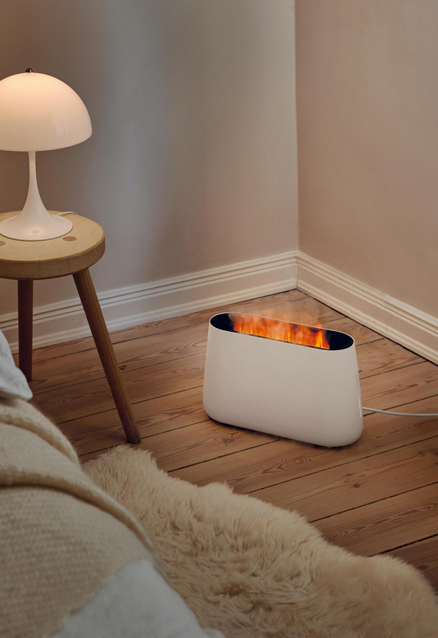 Biały kominek elektryczny z płomieniami, lampa i drewniany stołek w pokoju z beżowymi ścianami.