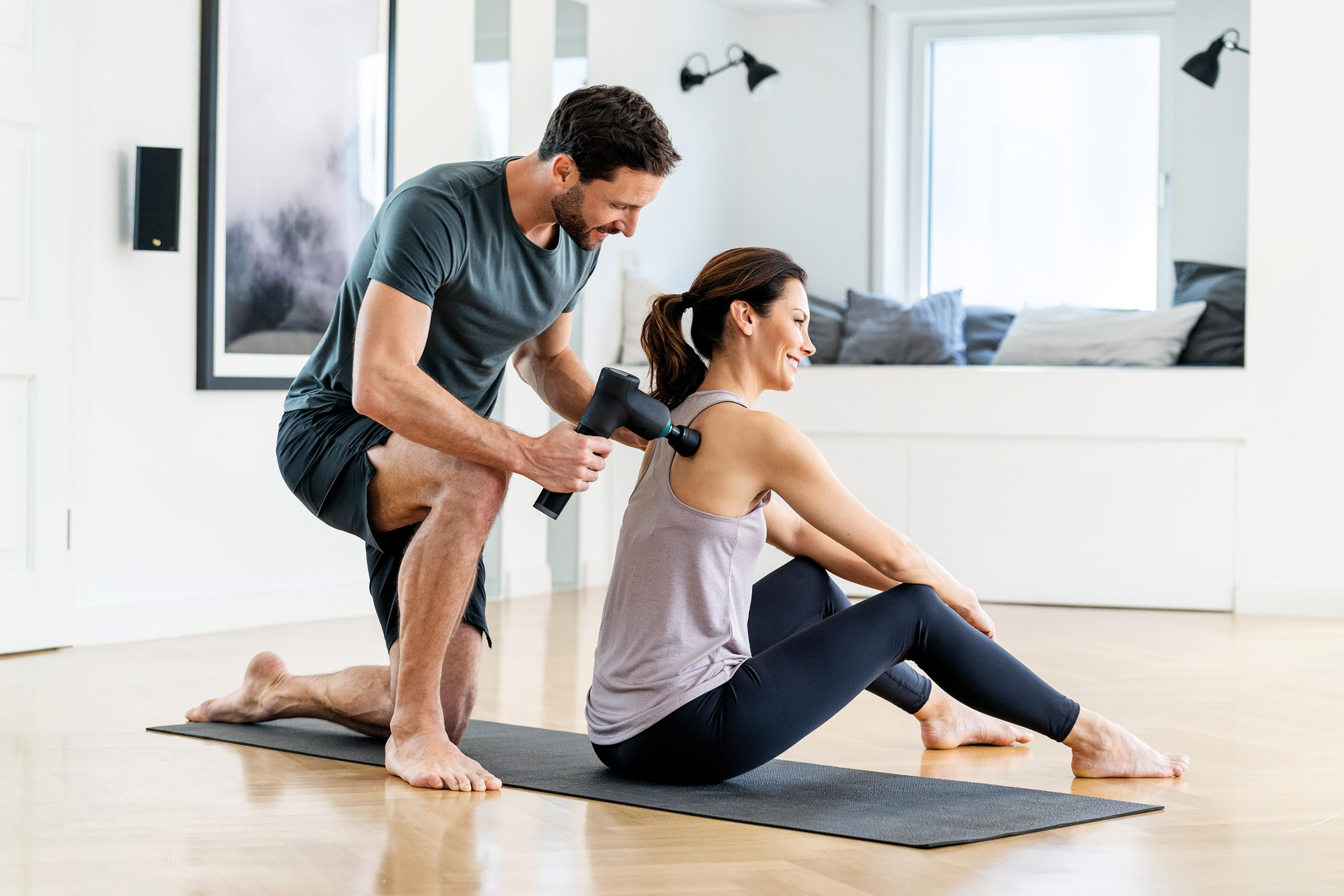 Ein Mann benutzt eine Massagepistole auf dem Rücken einer Frau, die auf einer Matte sitzt.
