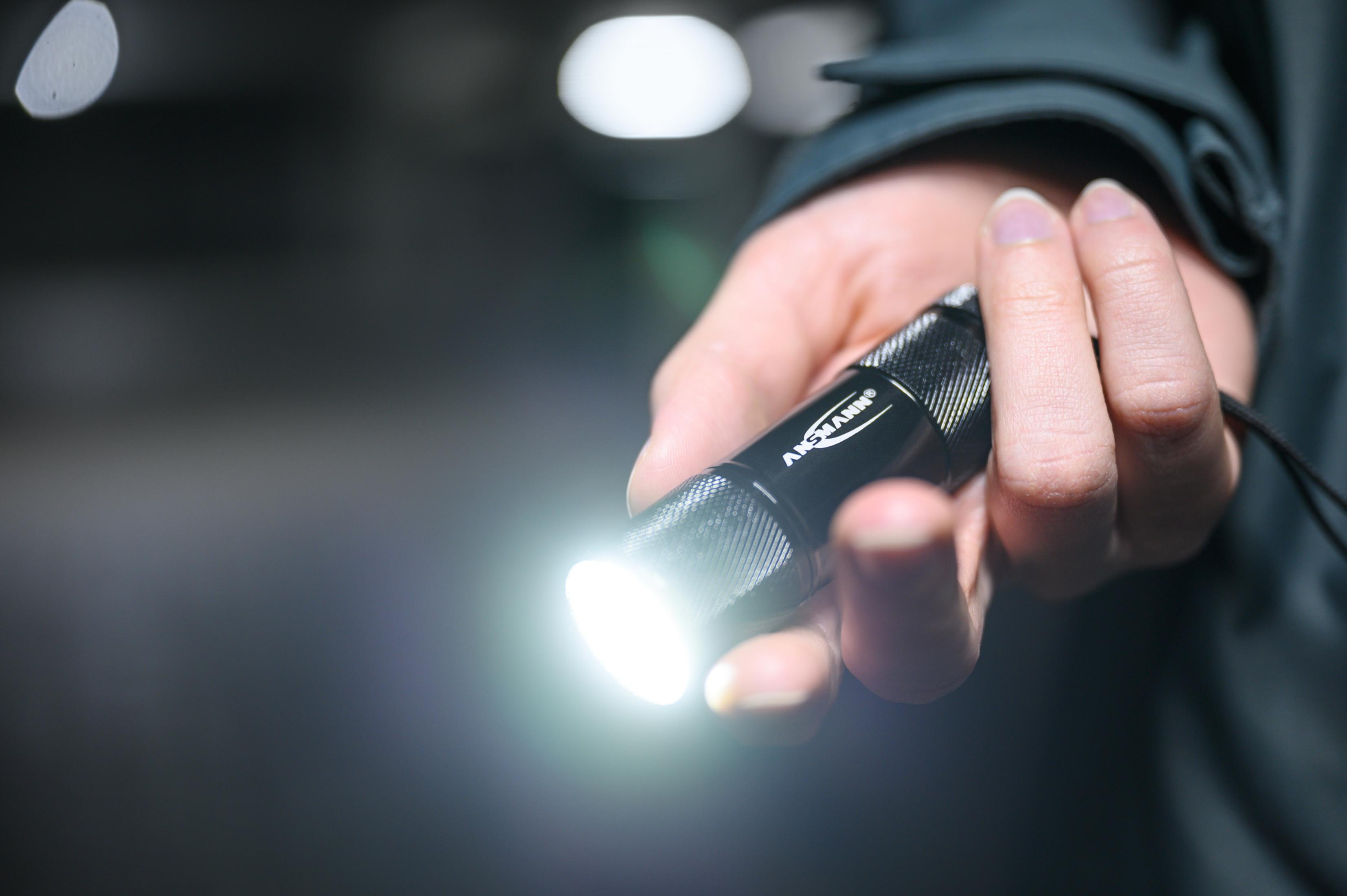 Hand hält eine schwarze Taschenlampe, die helles weißes Licht aussendet. Unscharfer Hintergrund.
