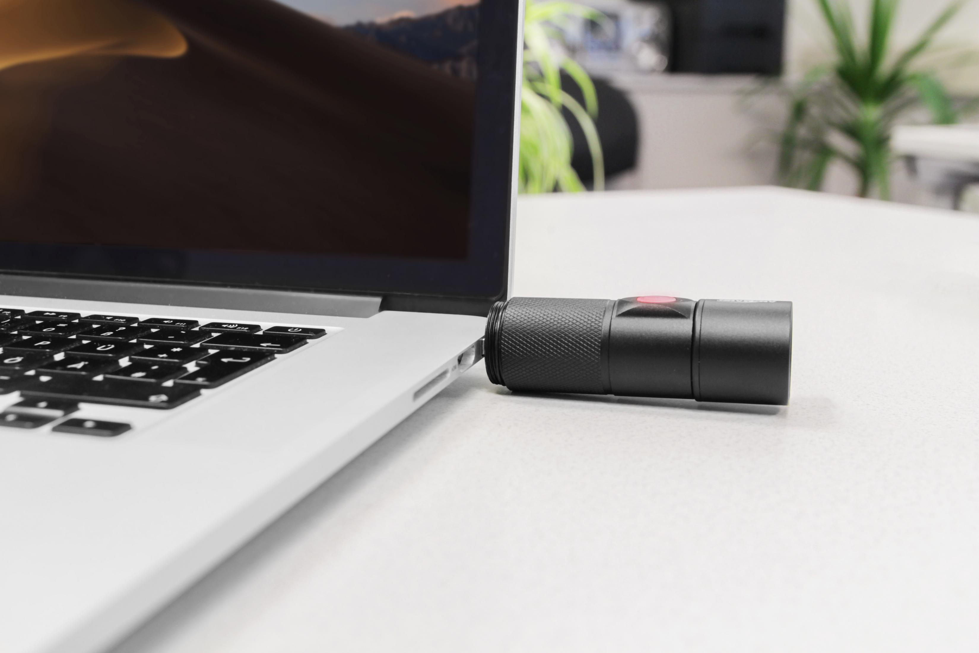 Czarne cylindryczne urządzenie jest podłączone do laptopa. Laptop na białym biurku. Widoczny czerwony przycisk.