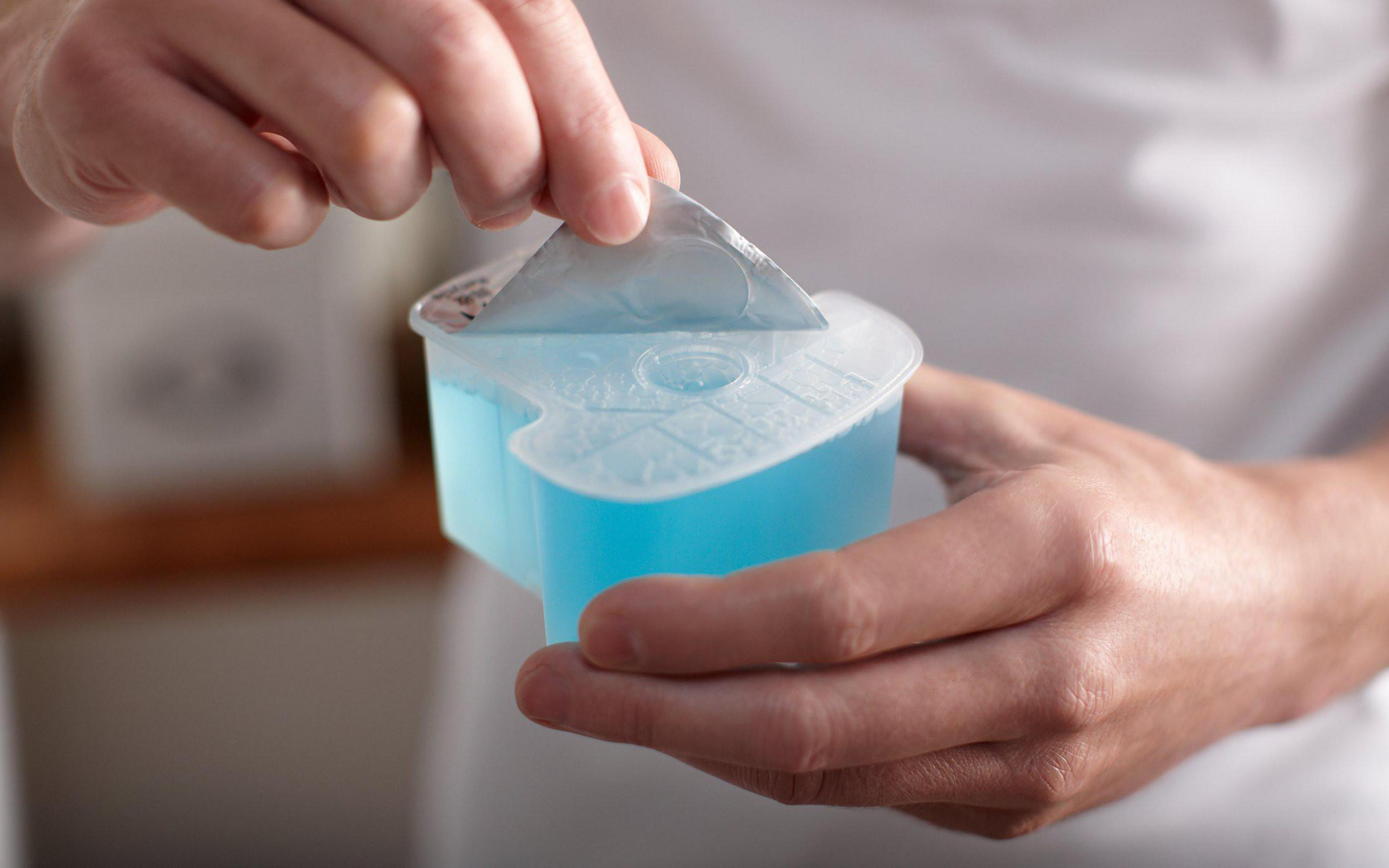 Manos abriendo un recipiente con líquido azul. El recipiente es blanco y tiene una tapa.