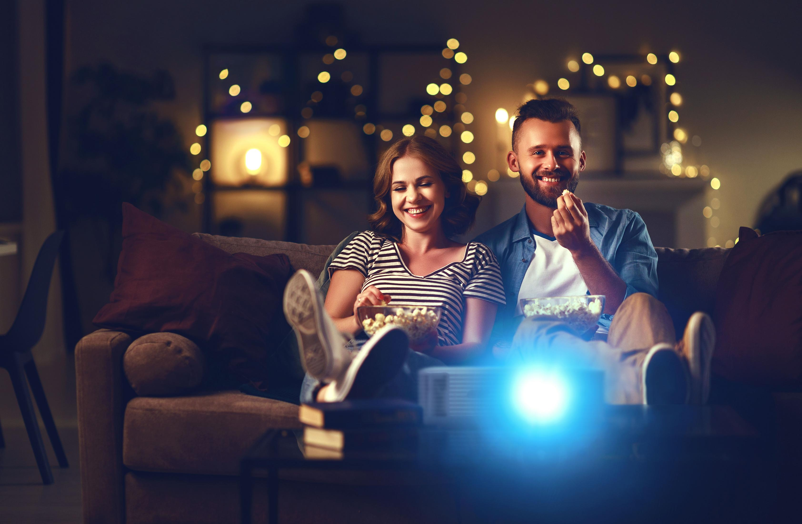 Pareja en sofá viendo película. Comen palomitas. Proyector al frente.