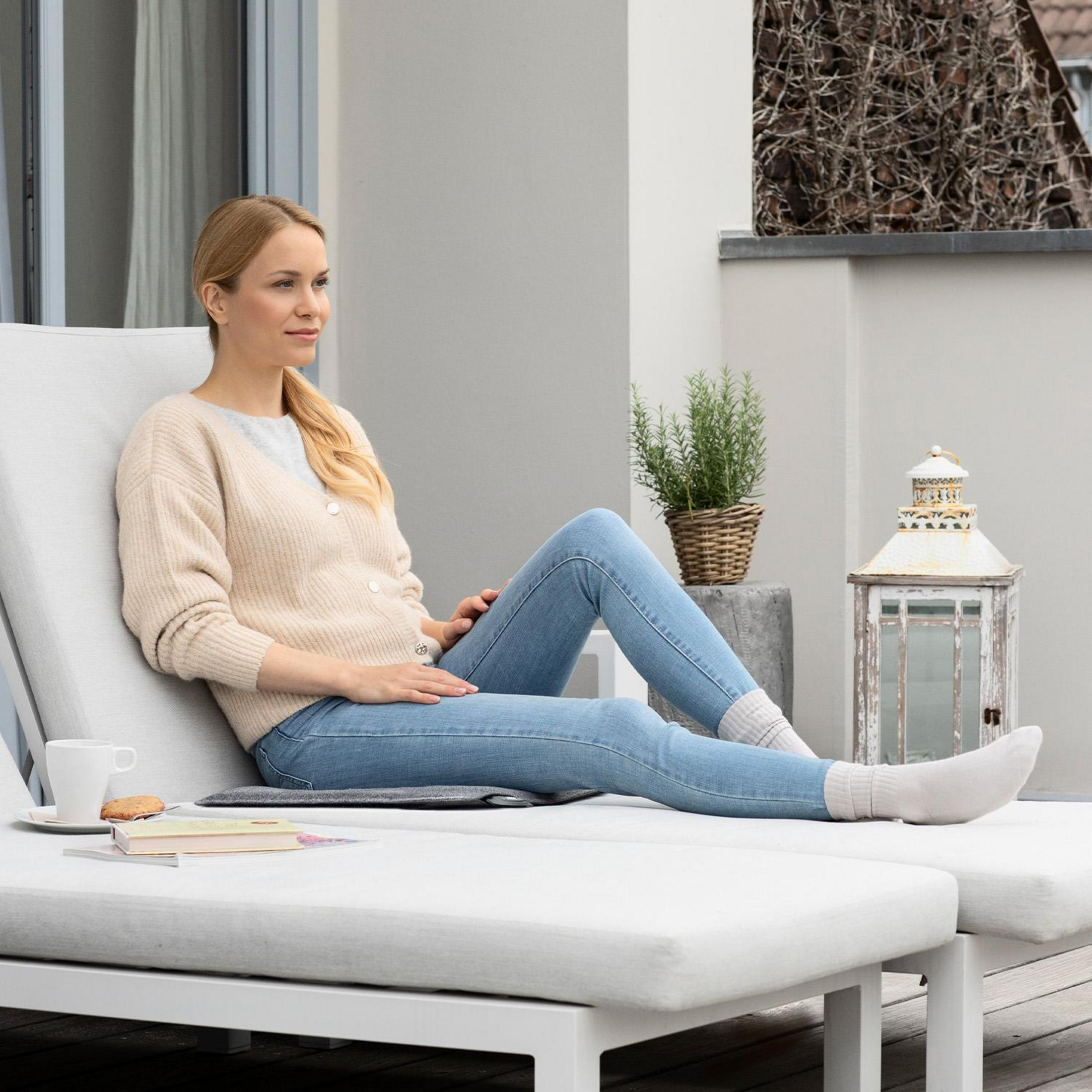 A woman relaxing on a white lounge chair outdoors, with a book and coffee cup nearby. Casual attire.