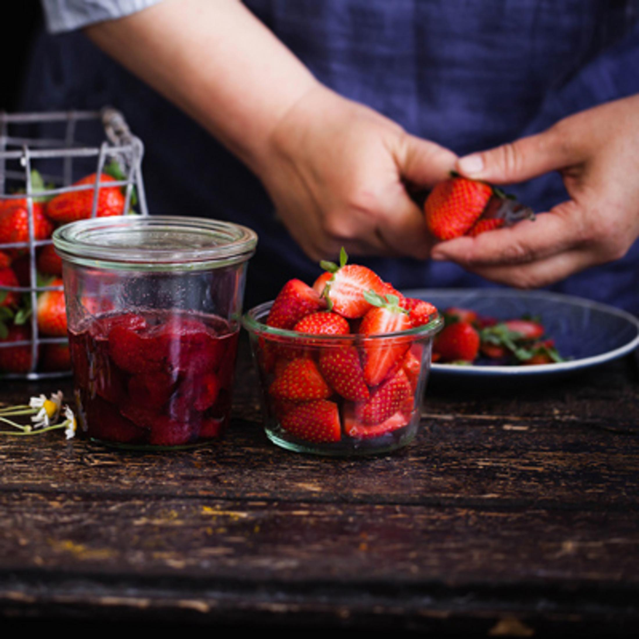 Una persona sostiene fresas sobre un tarro de cristal con mermelada. También hay un cuenco de fresas.
