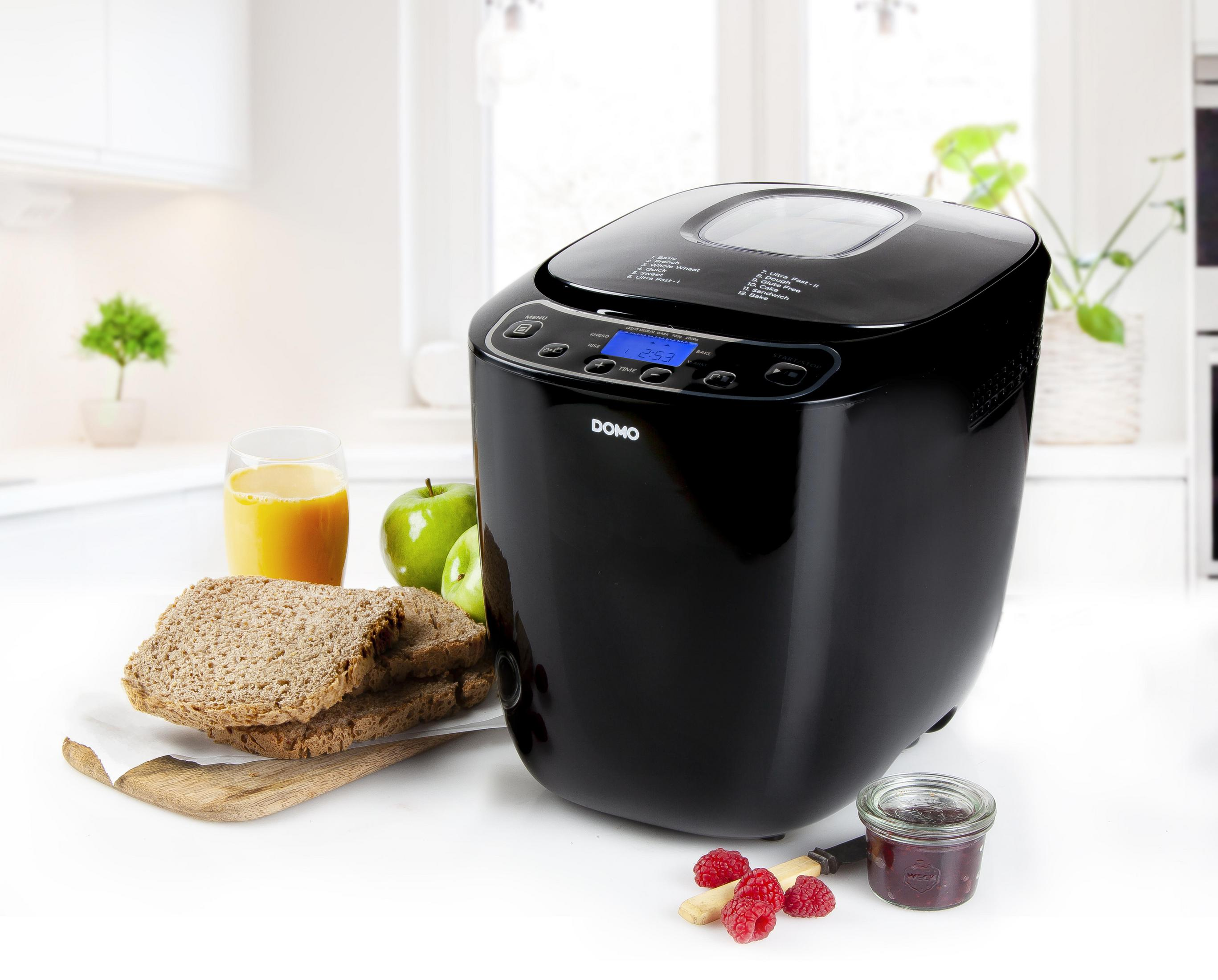 Schwarzer Brotbackautomat mit Display, Brot, Saft, Apfel, Marmelade und Himbeeren auf weißem Tisch.