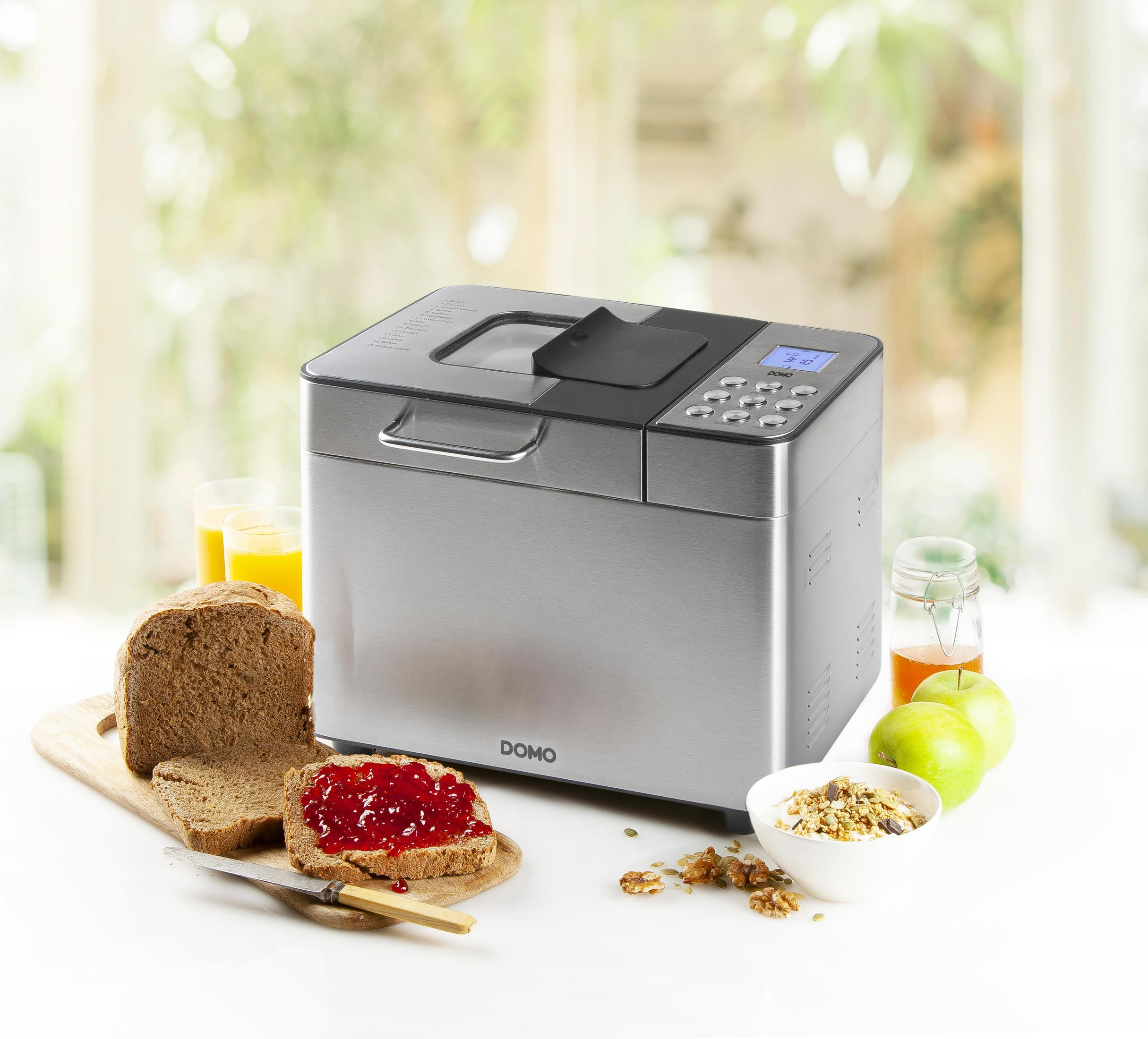 Brotbackmaschine mit Brot, Marmelade und anderen Lebensmitteln auf einem weißen Tisch.