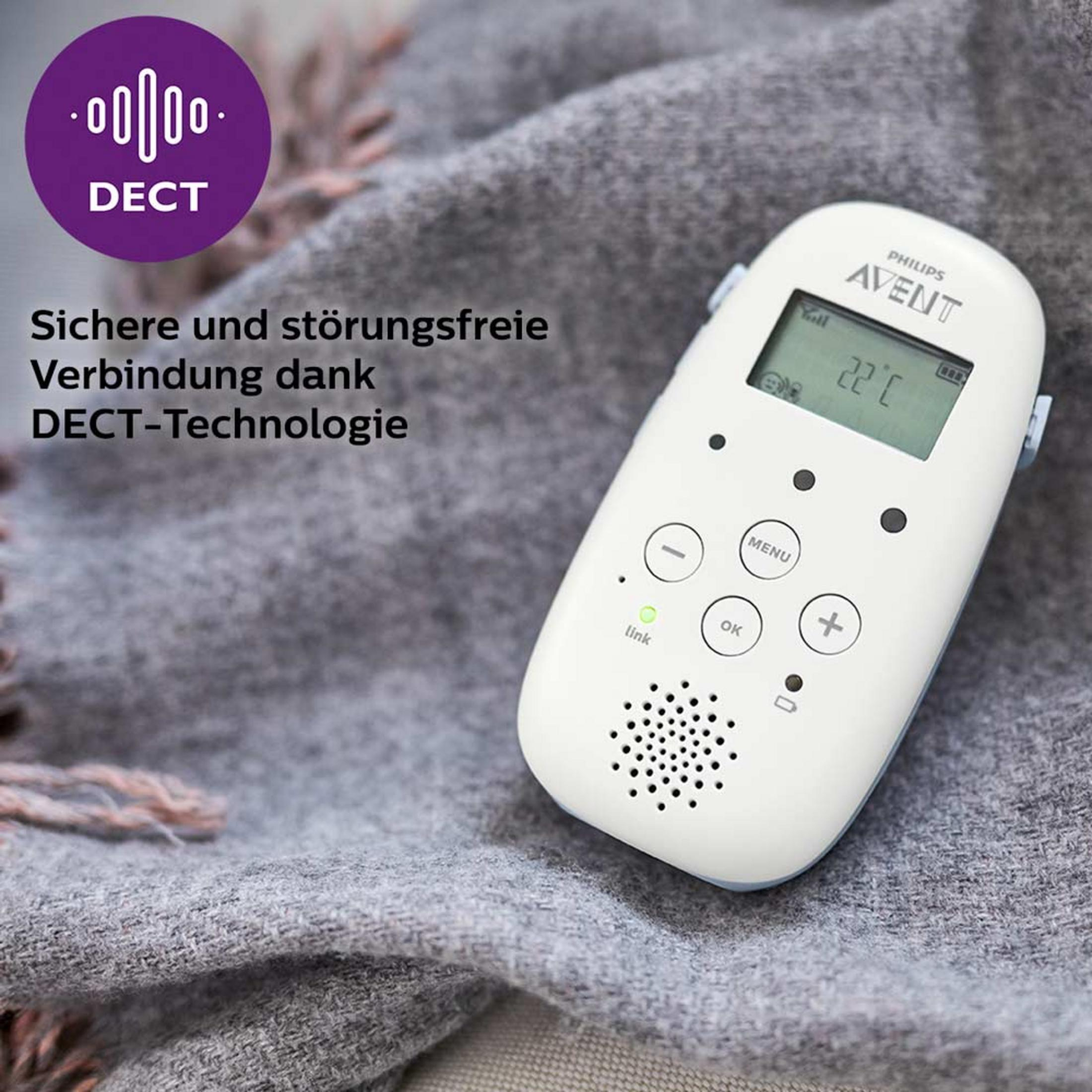 Ein Babyphone. Es hat Knöpfe und einen kleinen Bildschirm. Es liegt auf einer grauen Decke. Der Text ist auf Deutsch.