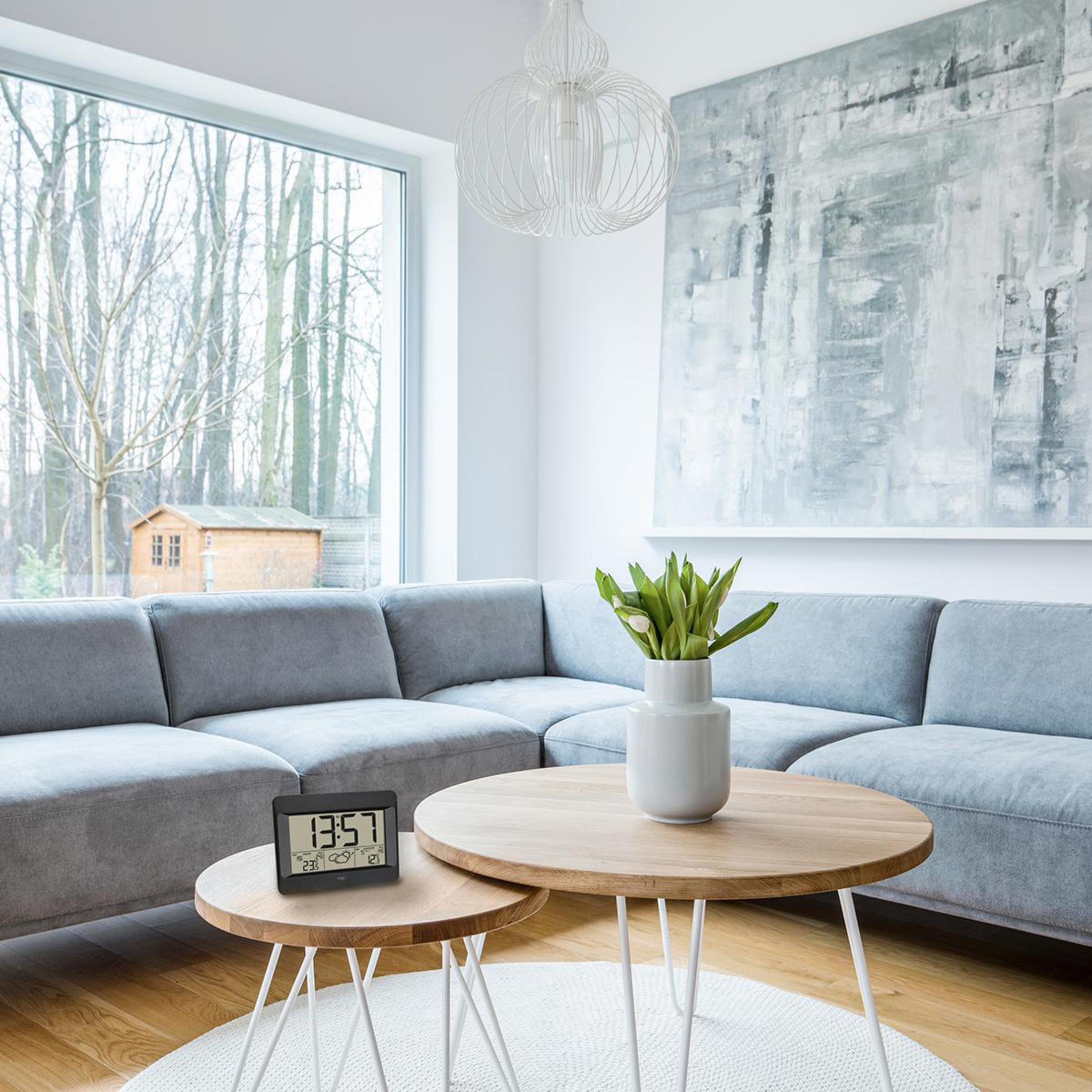 Wohnzimmer mit grauem Ecksofa, runden Holztischen und einer Digitaluhr.