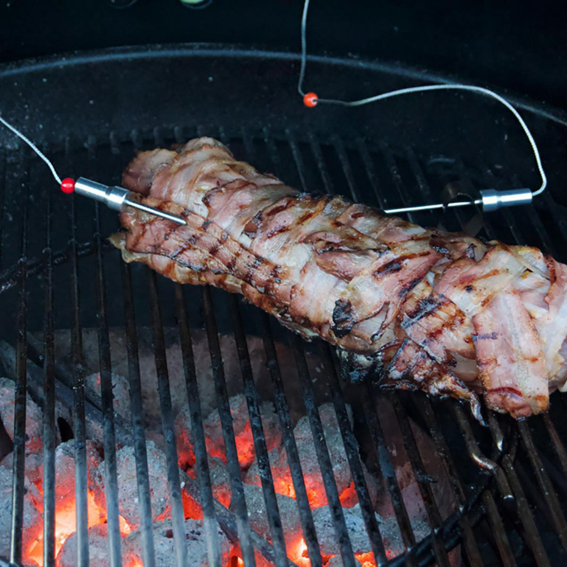 Mit Speck umwickeltes Fleisch auf einem Grill, mit Sonden eingesetzt. Der Grill ist schwarz, die Kohlen sind angezündet.