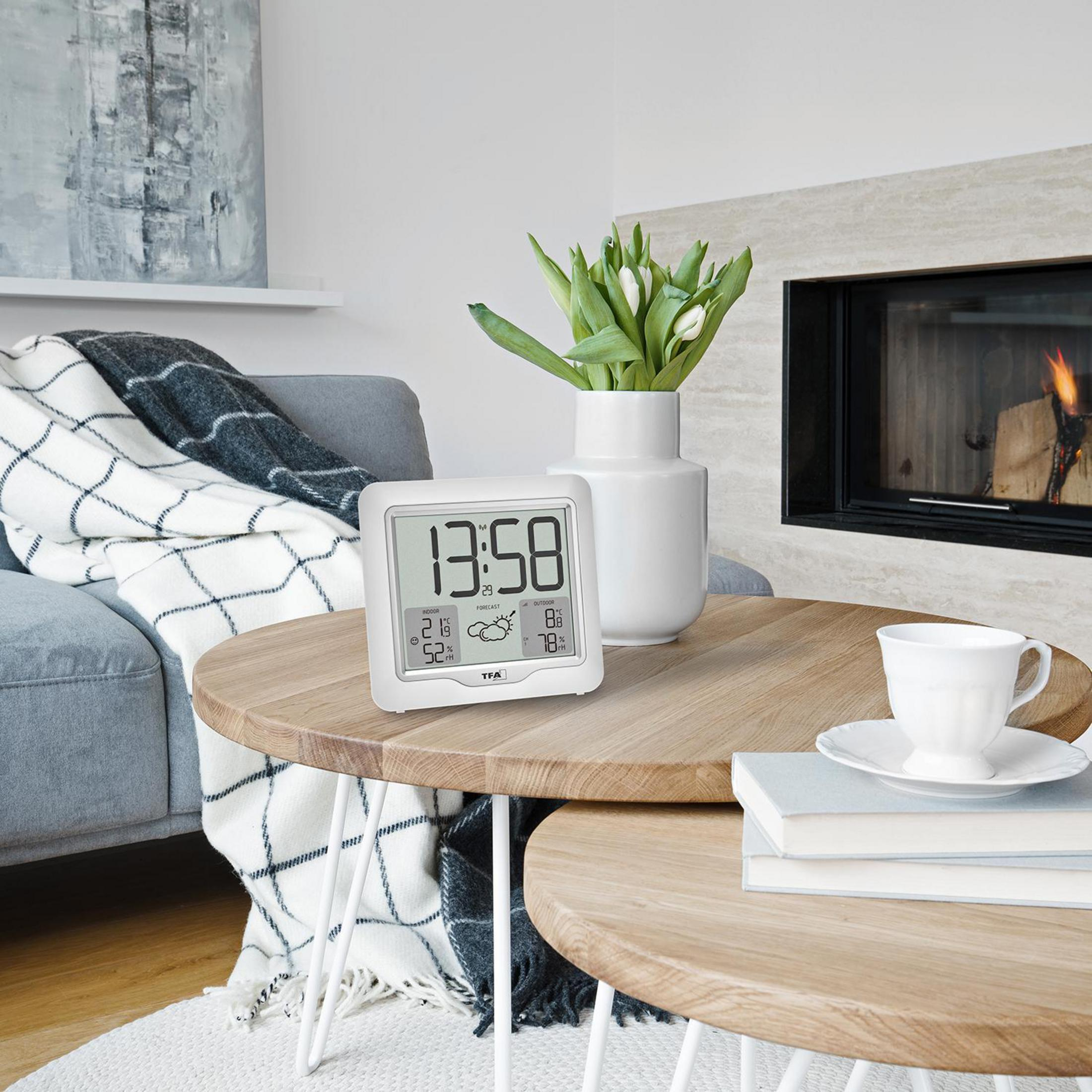 Eine weiße YTA-Wetterstation auf einem Holztisch, mit einer Tasse und einer Blumenvase. Kamin im Hintergrund.