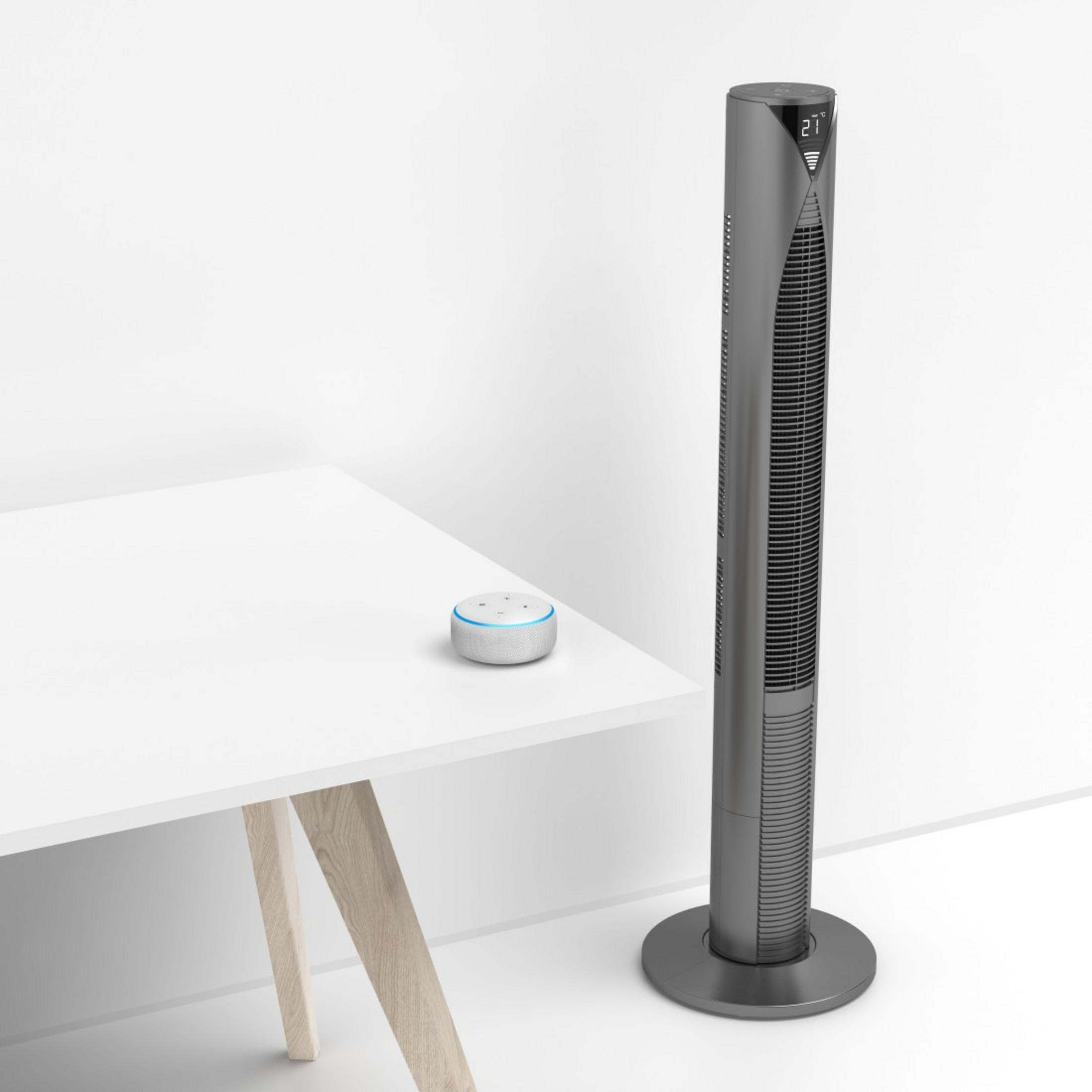 Ventilador gris alto junto a un escritorio blanco con un altavoz inteligente. Fondo blanco.