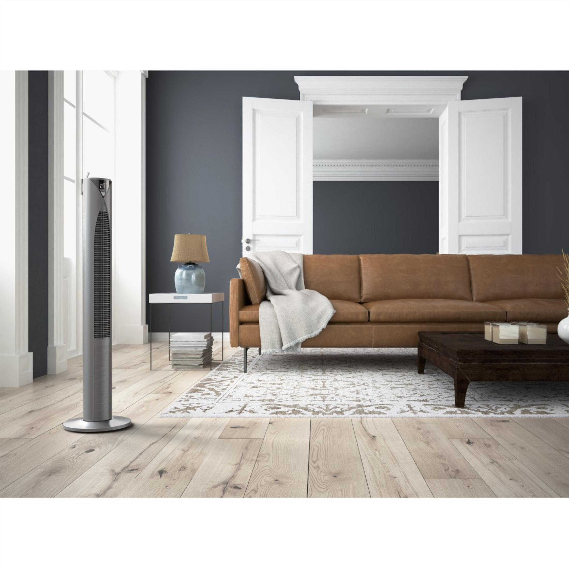Un ventilador alto y plateado está en un piso de madera. Un sofá marrón, una lámpara y una alfombra están detrás.
