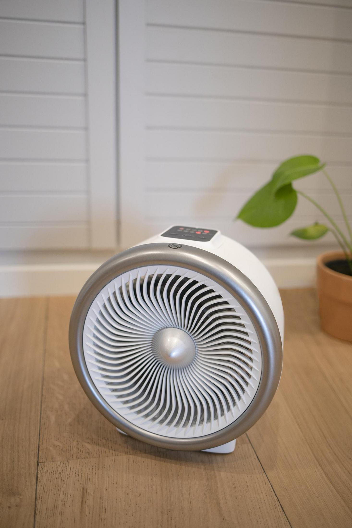 Ventilatore bianco con un anello argentato, su un pavimento in legno. Visibile anche una pianta verde in vaso.