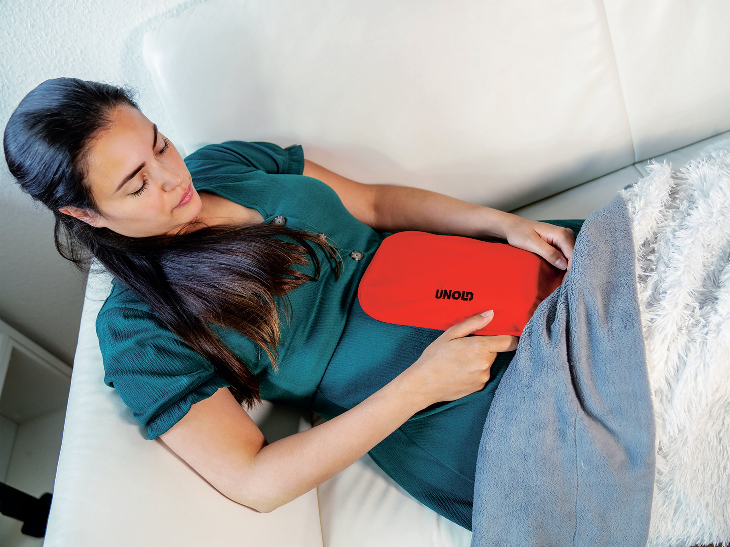 Kobieta leżąca na kanapie z czerwoną butelką z gorącą wodą na brzuchu.