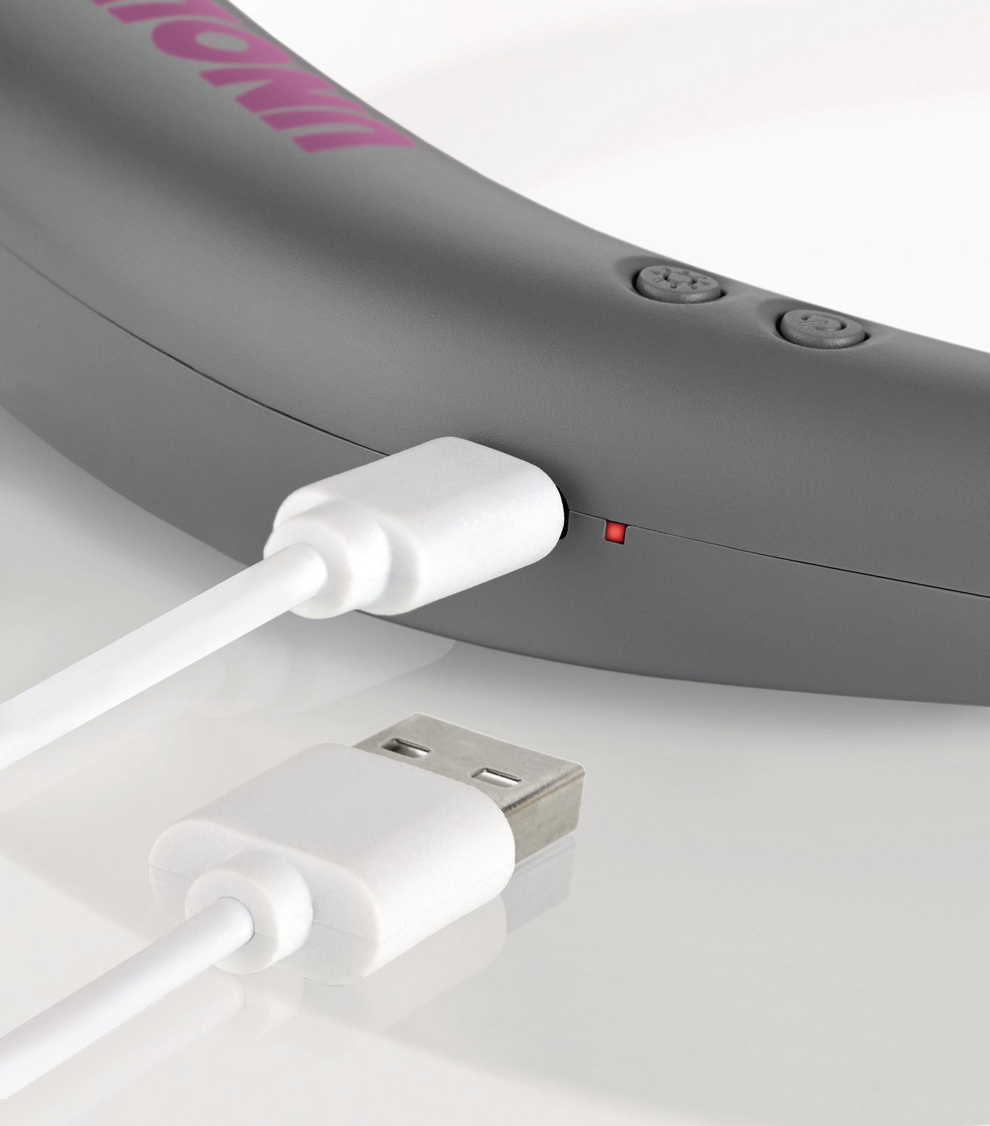 Szare urządzenie z białym kablem ładującym; czerwone światło wskazuje ładowanie.