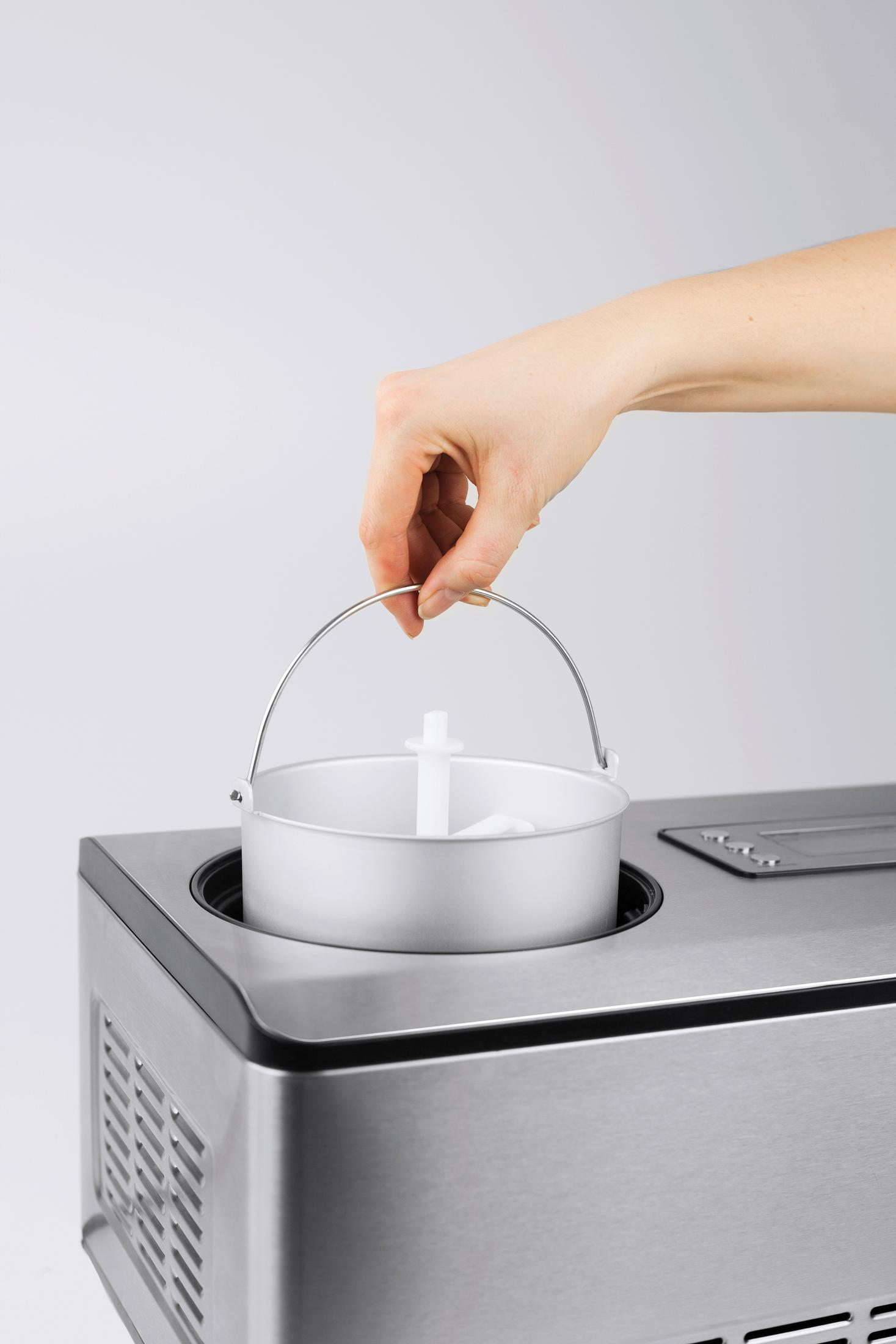 Eine Hand hält einen Metalleimer mit einem weißen Kunststoffteil darin, über einer Edelstahlmaschine. Weißer Hintergrund.