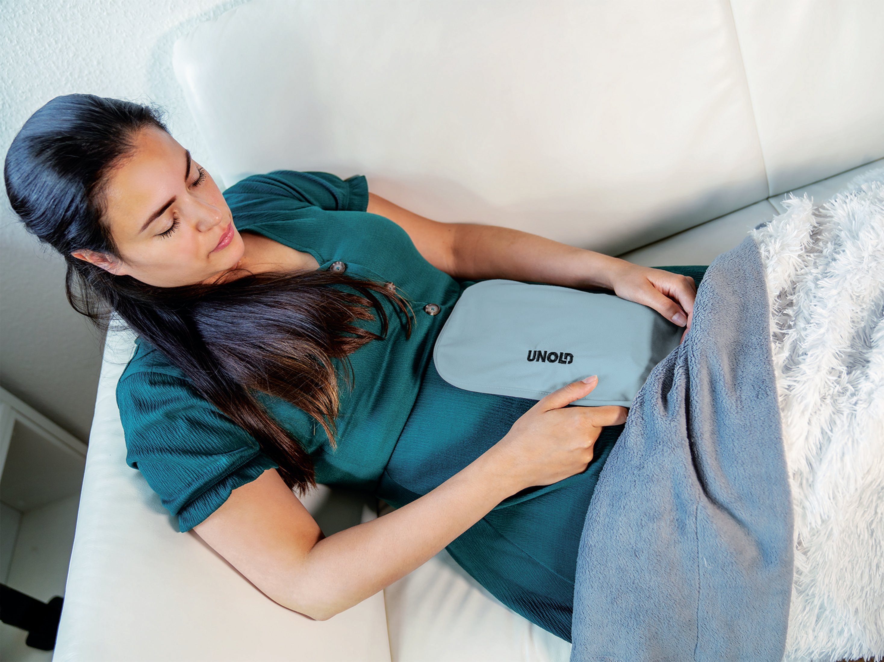 Kobieta odpoczywająca na białej kanapie z termoforem na brzuchu.