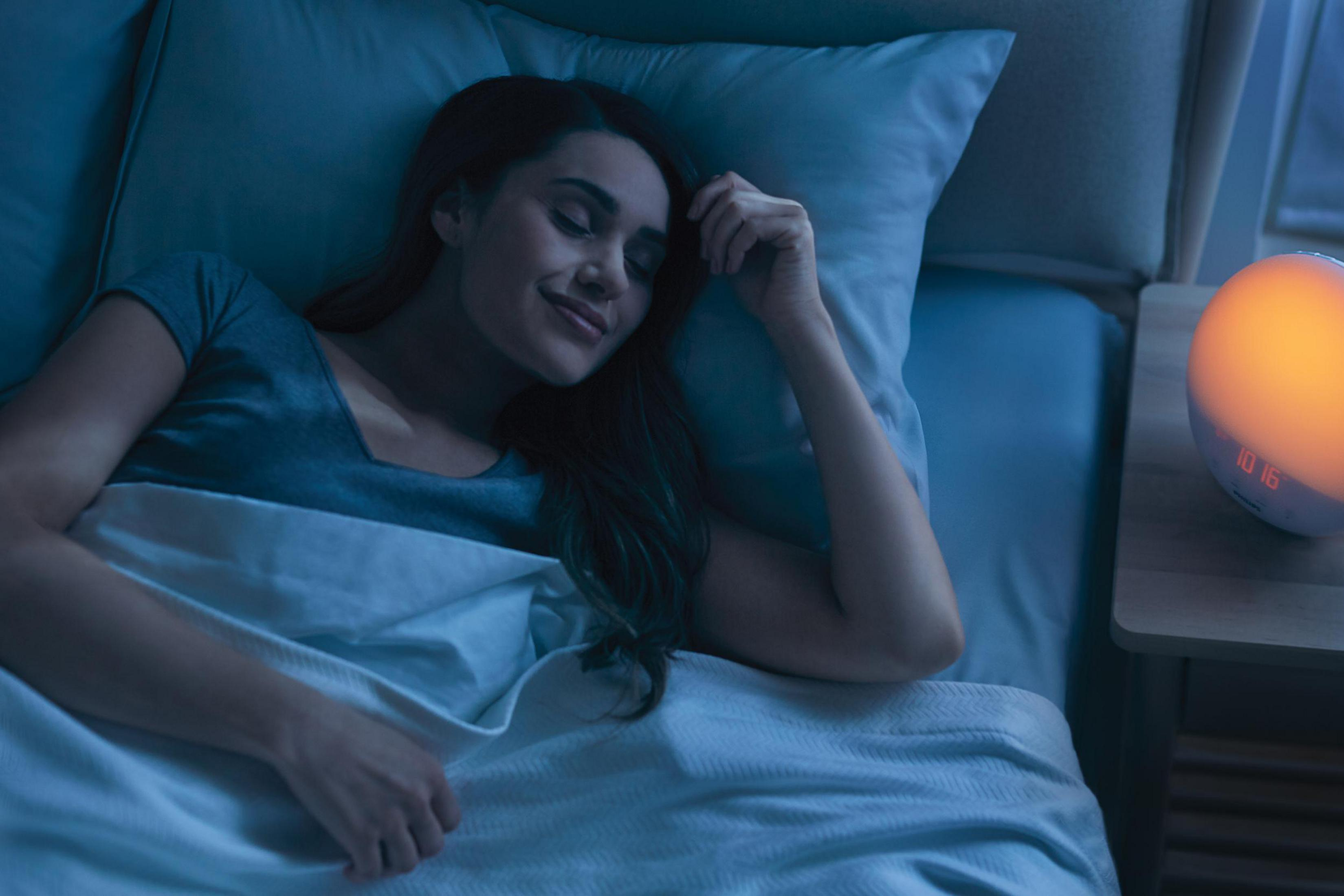 Frau schläft im Bett mit einem Nachttischuhr. Bett ist weiß und blau. Dunkler Raum.