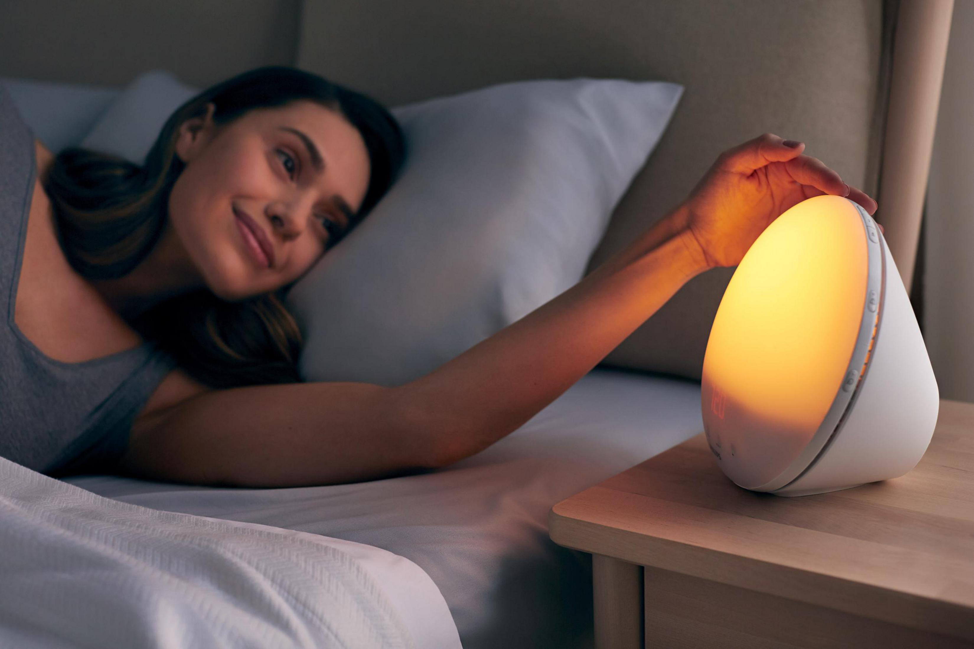 Frau im Bett greift nach einem weißen Wecker mit gelbem Licht.