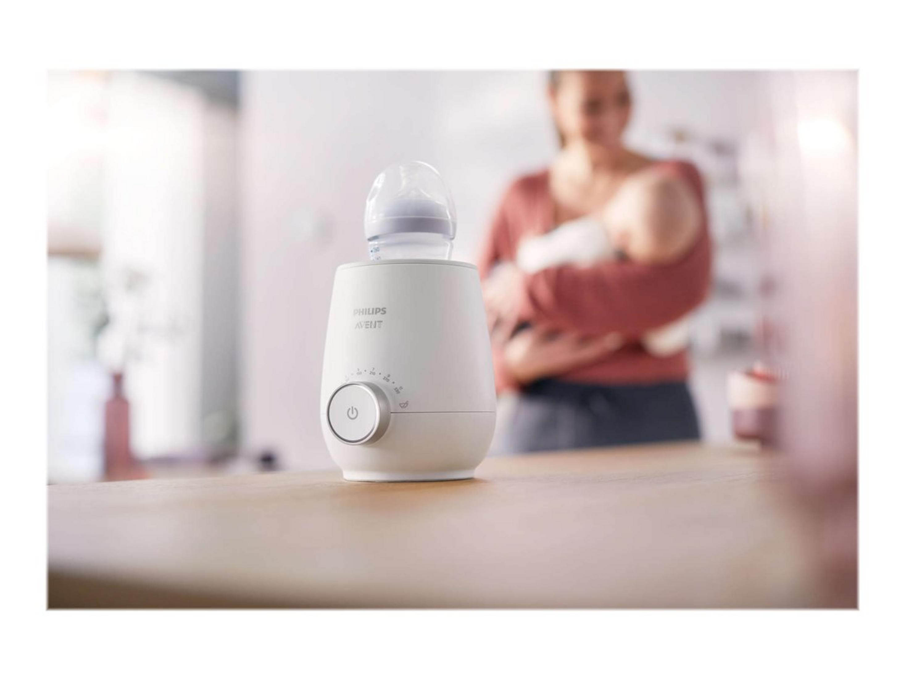 Weißer Philips Avent Flaschenwärmer mit Flasche, im Hintergrund Frau mit Baby.