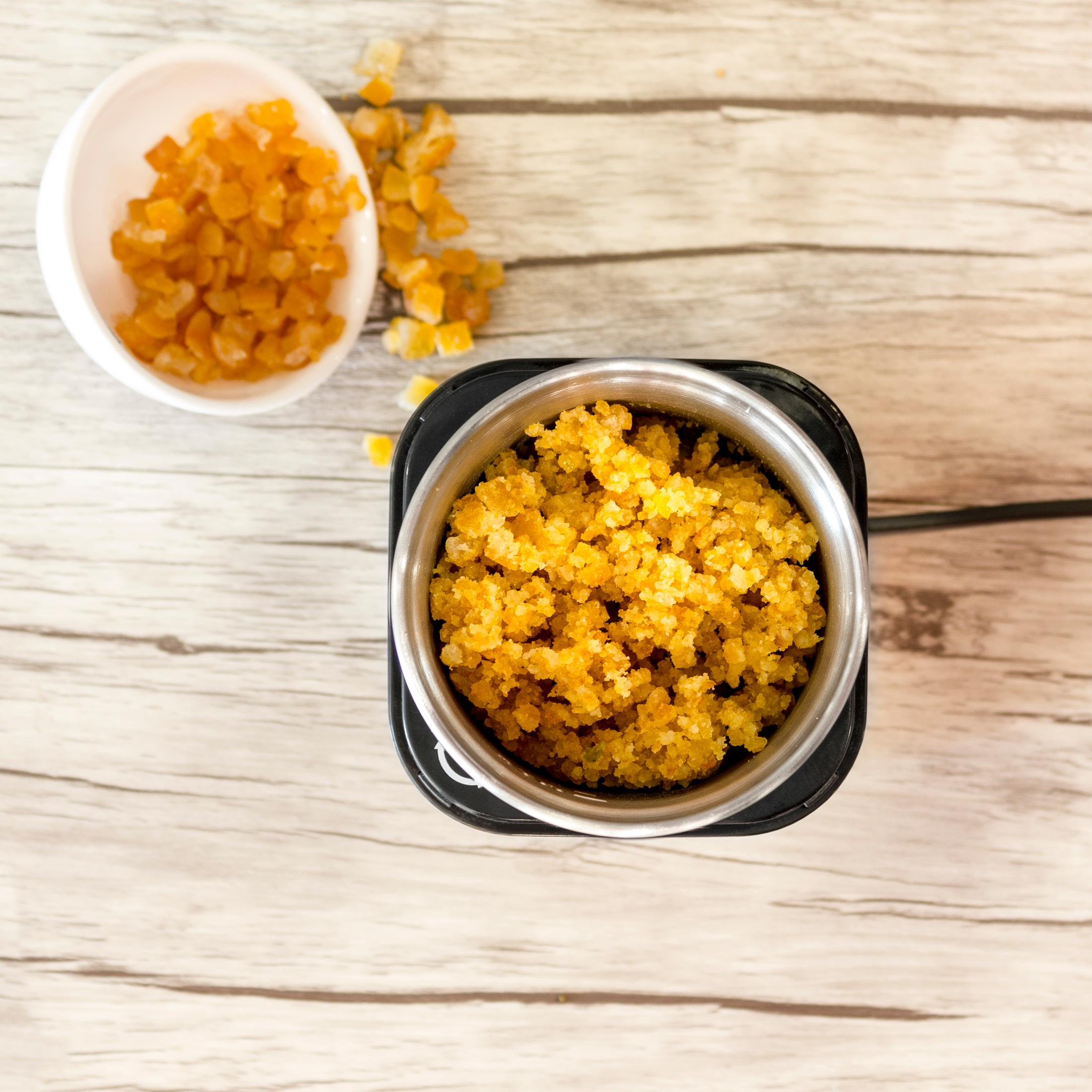 Widok z góry na młynek do kawy z pomarańczowymi granulkami i białą miseczką z granulkami.