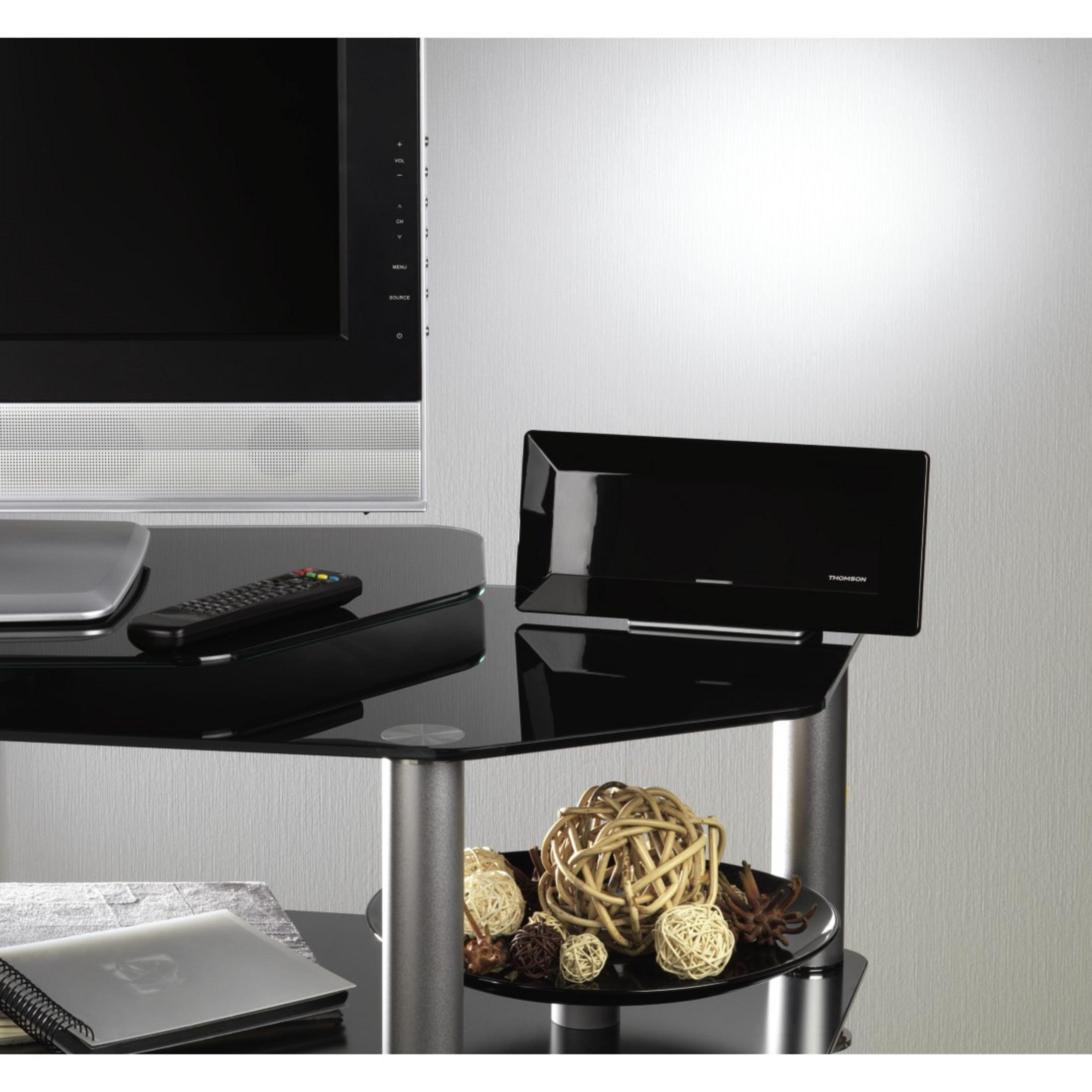 Czarny telewizor na szklanym stoliku z głośnikiem, pilotem i dekoracjami. Jasna ściana.