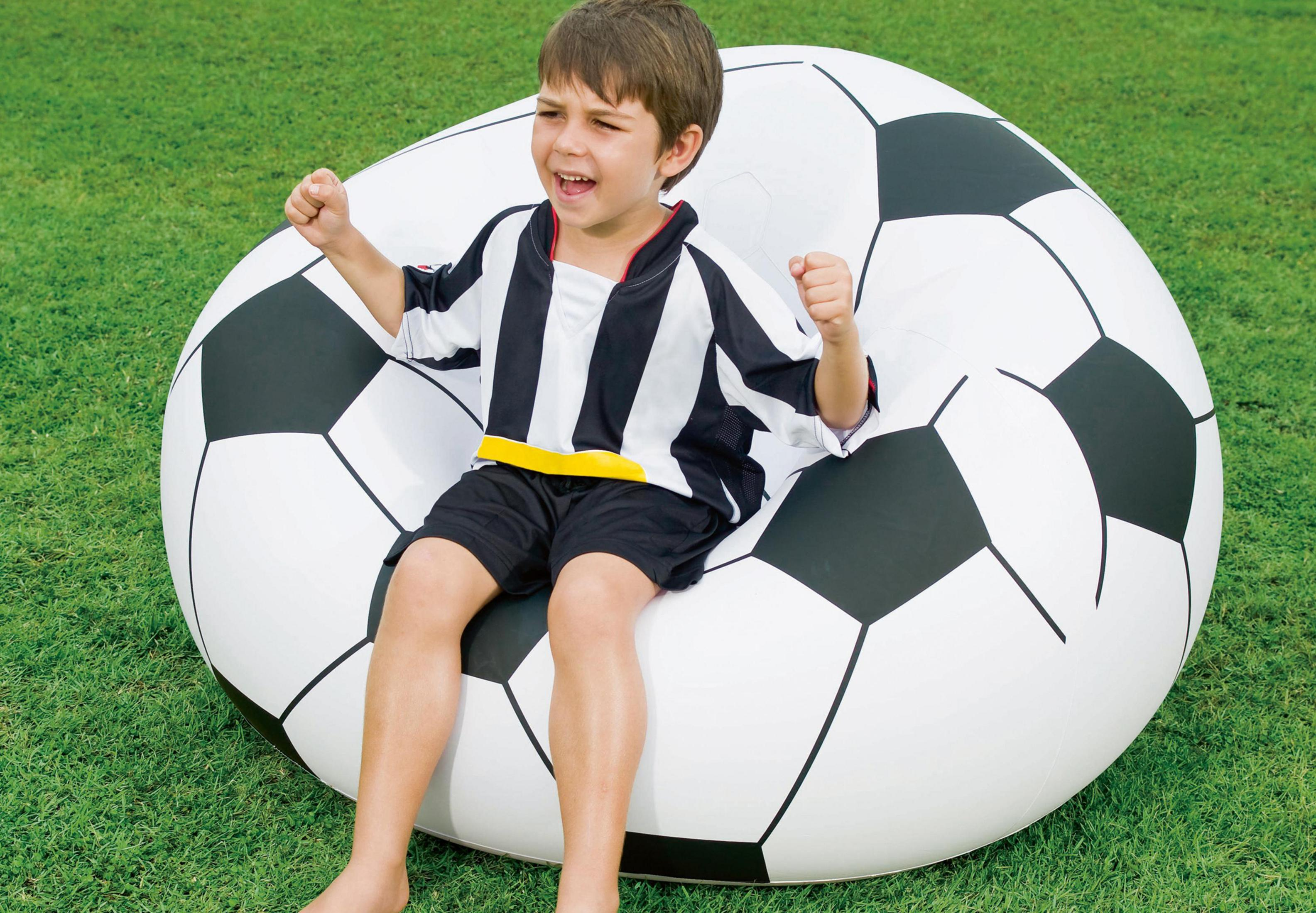 Ein Junge im Fußballtrikot sitzt auf einem aufblasbaren Fußball auf grünem Gras und lächelt.