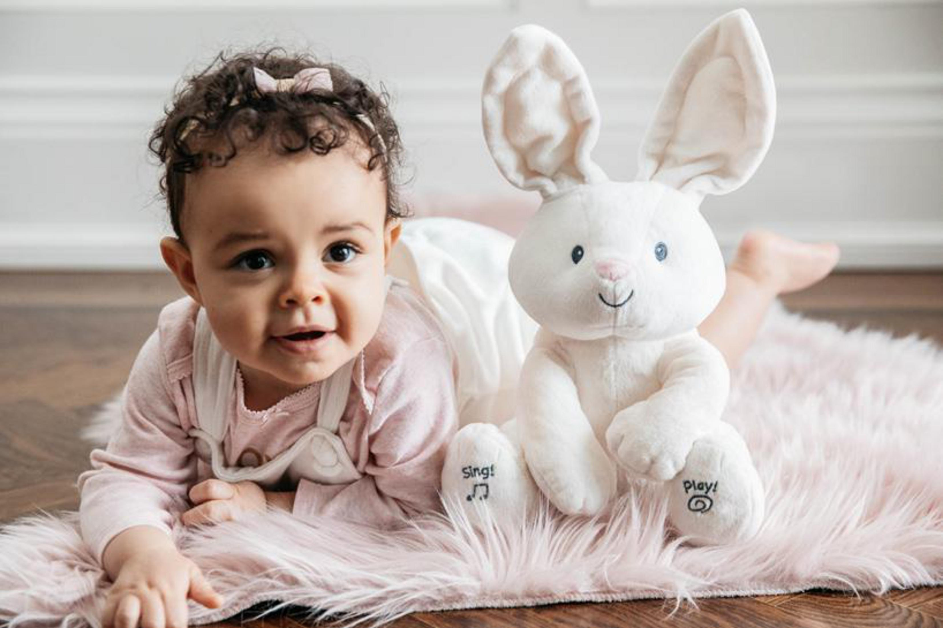 Baby-Mädchen liegt auf rosa Fell, lächelt, mit einem weißen Plüsch-Hase-Spielzeug.