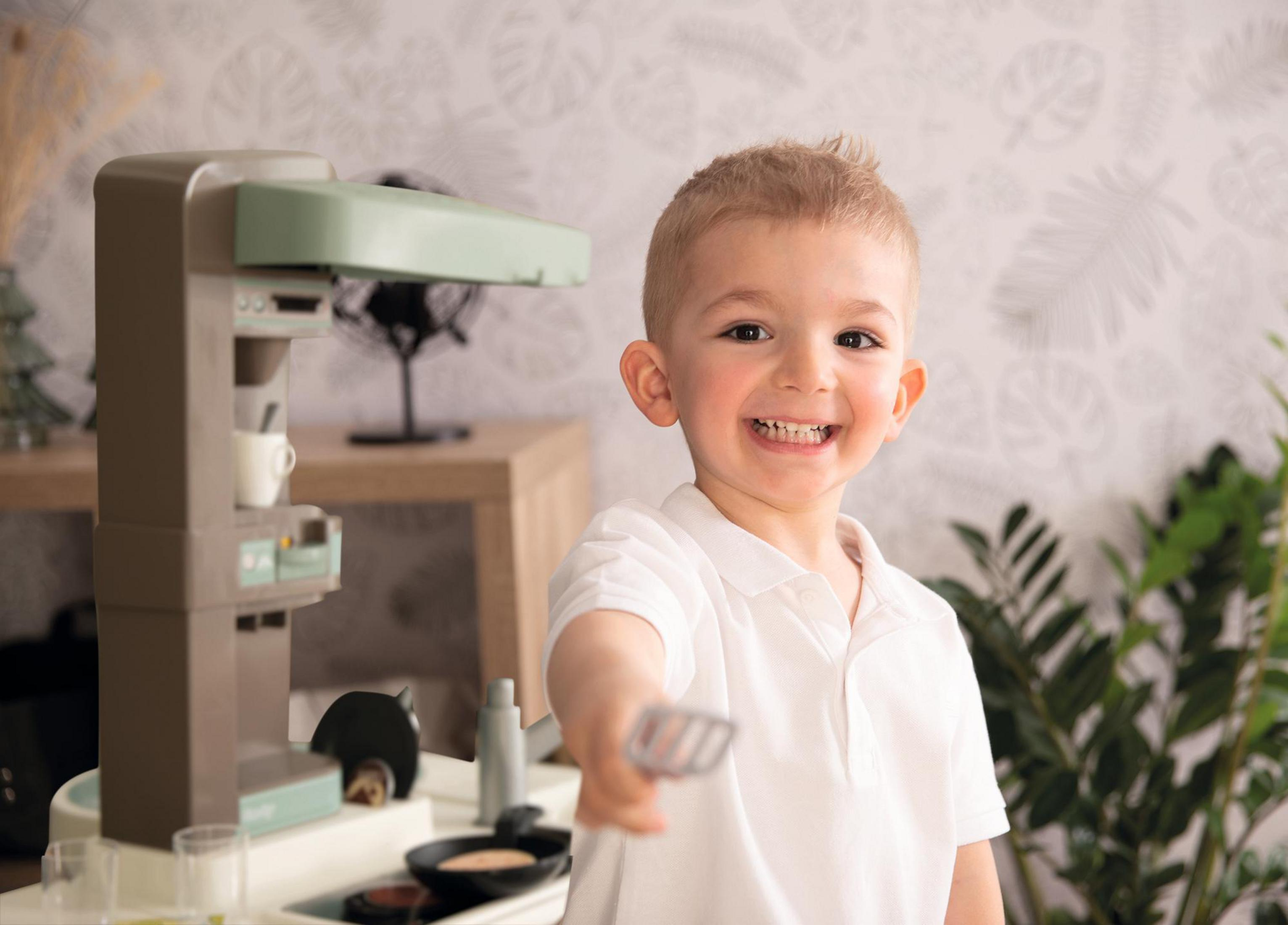 Mały chłopiec uśmiecha się, trzymając szpatułkę. Za nim kuchnia zabawka z ekspresem do kawy i wentylatorem.