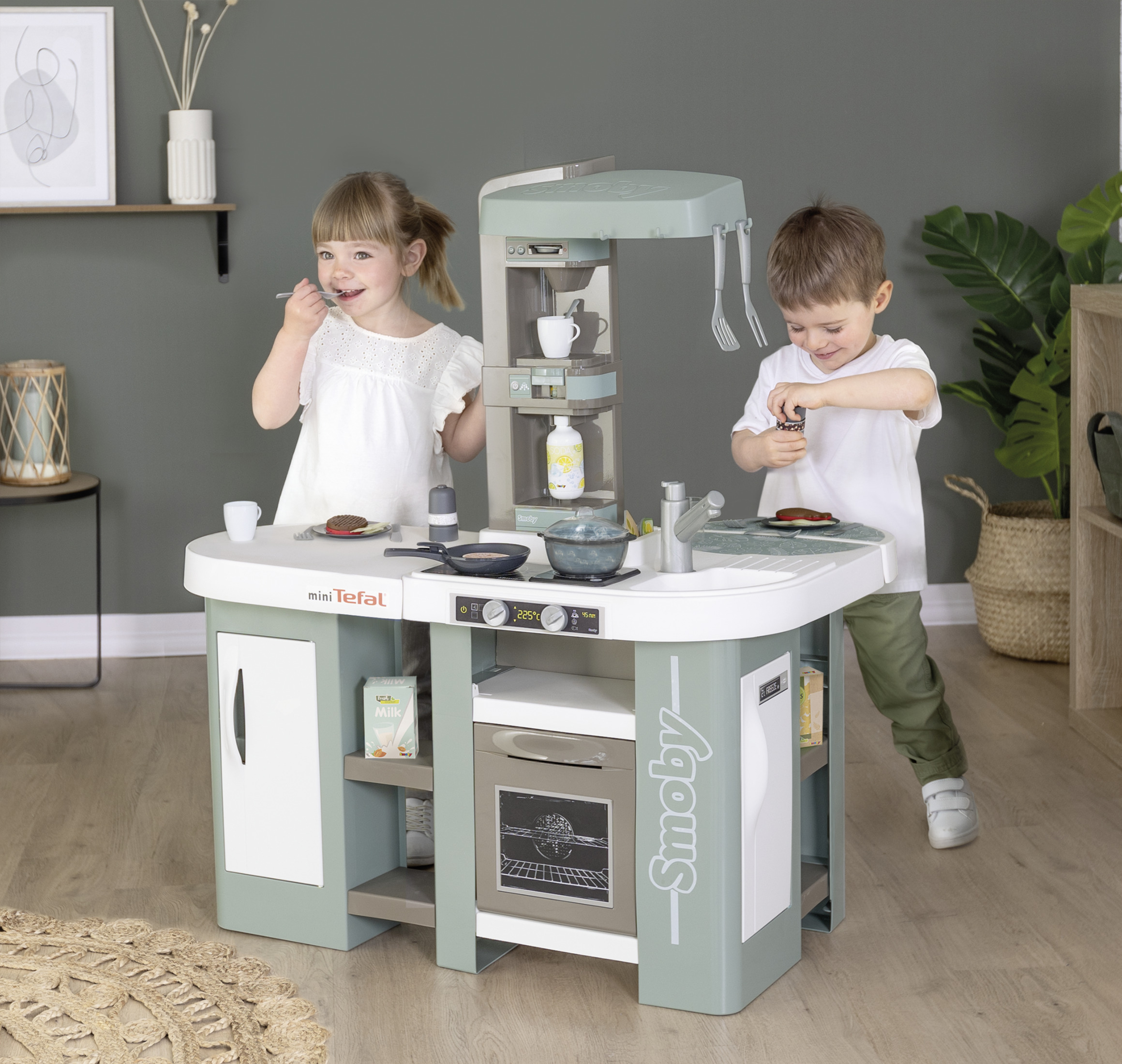 Dwoje dzieci bawiących się w kuchni dla dzieci. Kuchnia jest jasnozielona i biała. Drewniana podłoga.
