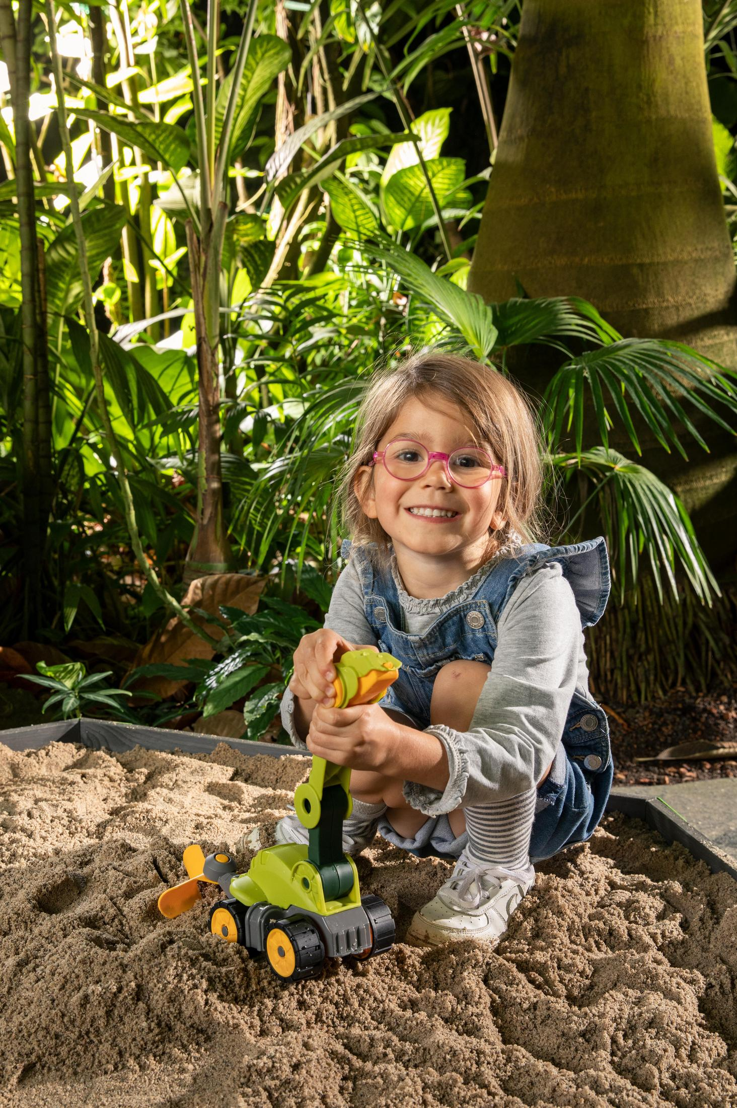 Ein junges Mädchen spielt mit einem Spielzeugbagger in einem Sandkasten und lächelt in die Kamera.