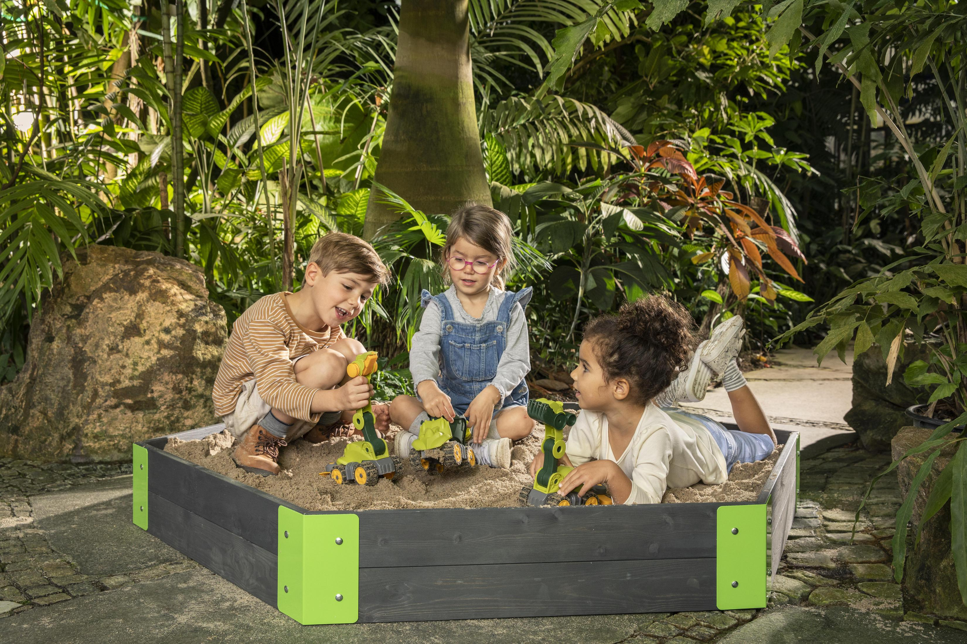 Drei Kinder spielen mit Spielzeug-LKWs in einem Sandkasten. Üppige grüne Pflanzen im Hintergrund. Sonniger Tag.