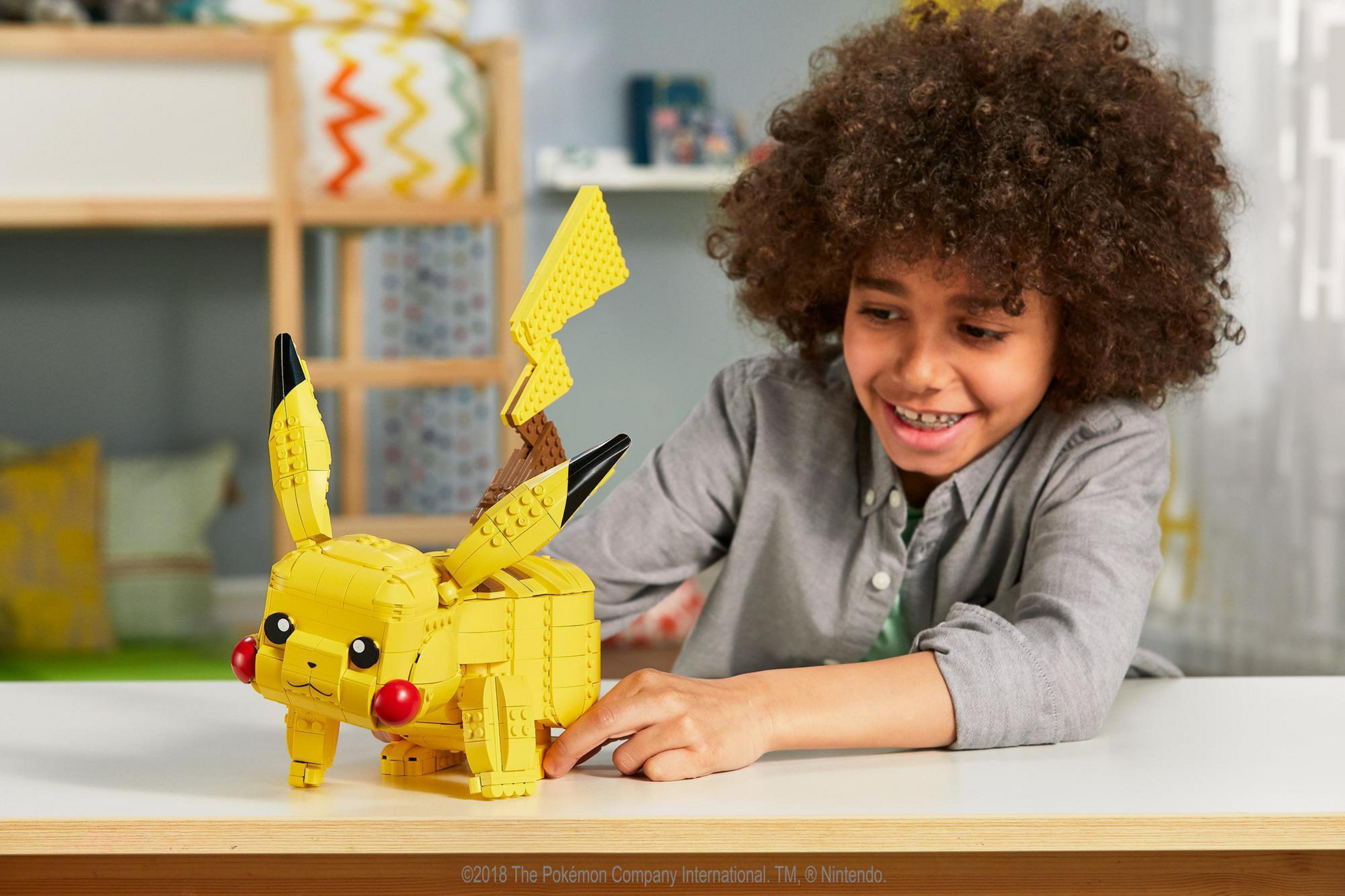 Niño sonriendo, construyendo un set Lego de Pikachu amarillo. Mesa blanca, estantería en el fondo.
